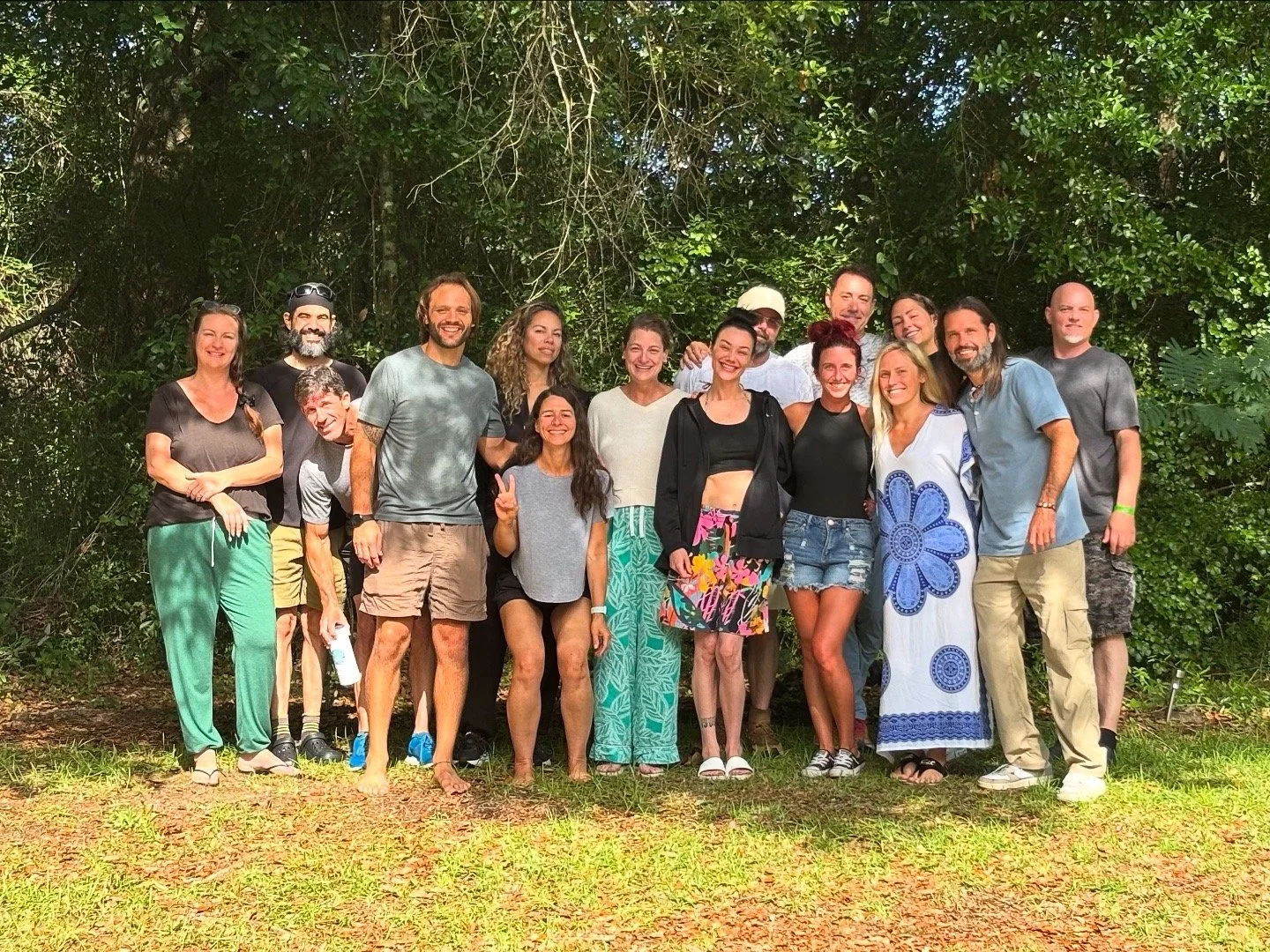 Group of 16 people standing outdoors in front of green foliage, smiling and posing for a photo.