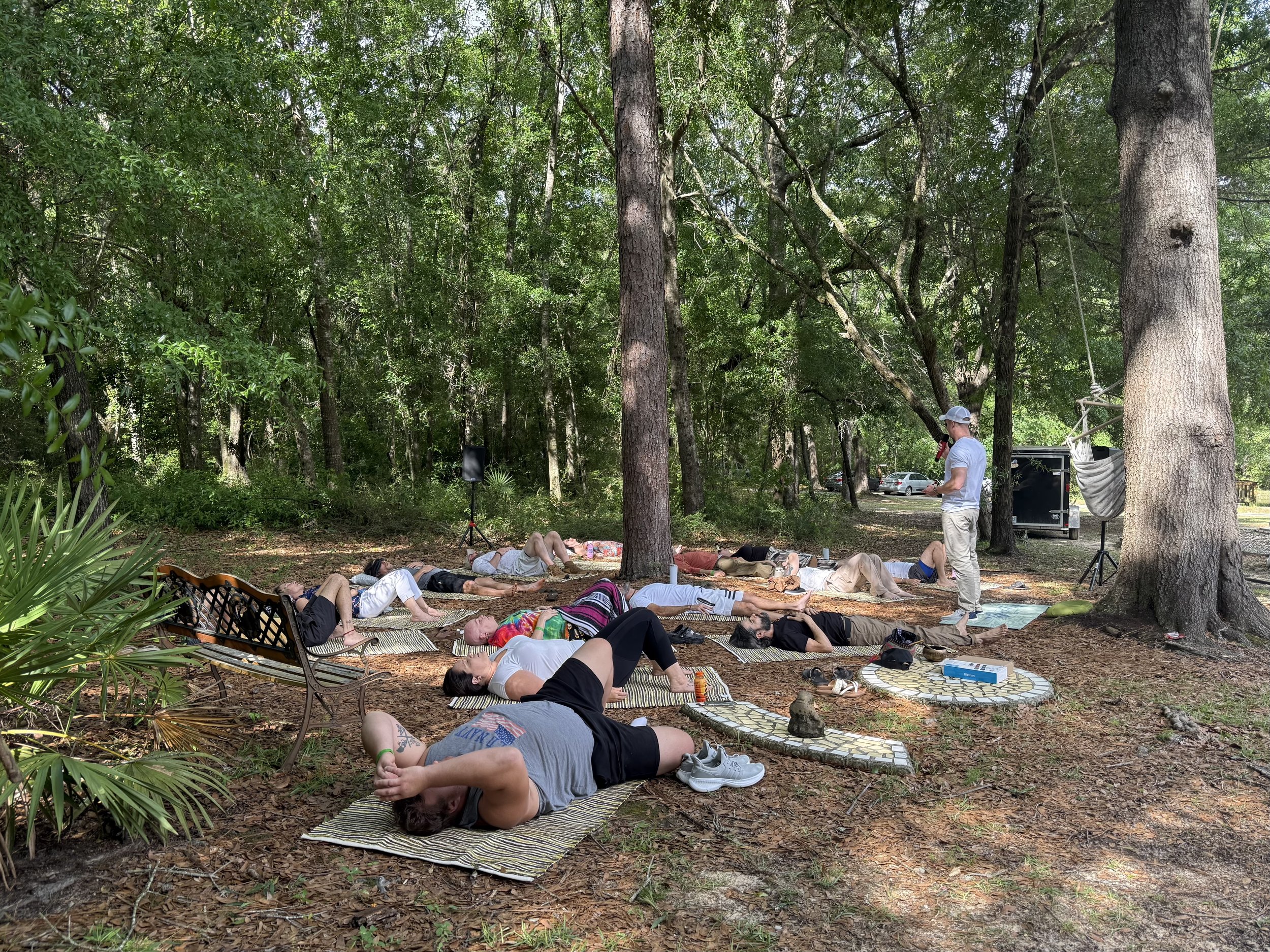 A group of people lying on mats in a wooded outdoor area, participating in a meditation or relaxation session with a person standing nearby.
