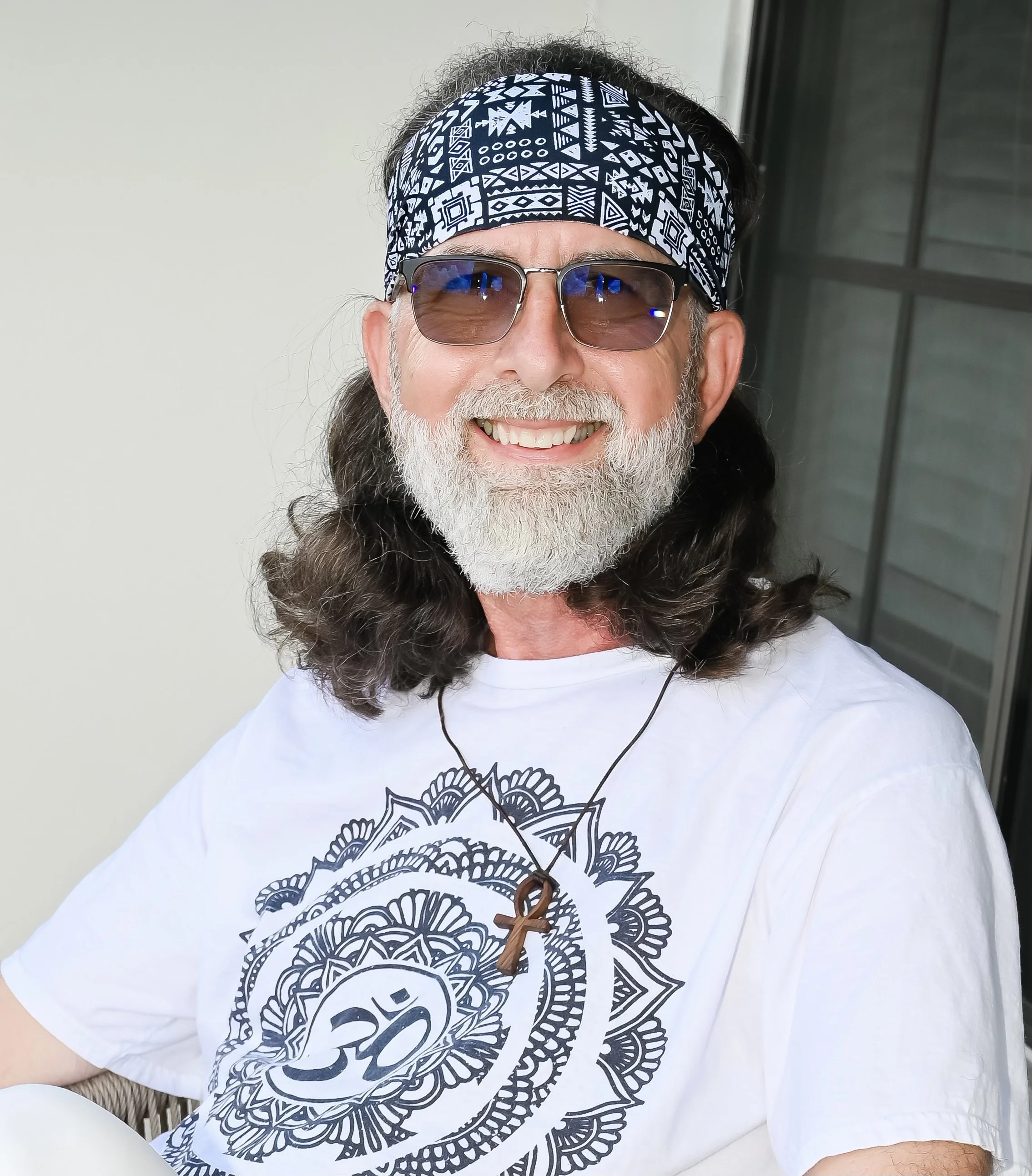 Smiling middle-aged man with long hair and beard wearing sunglasses, a bandana, a white T-shirt with a black mandala design, and a necklace with an ankh pendant, sitting outside near a window.