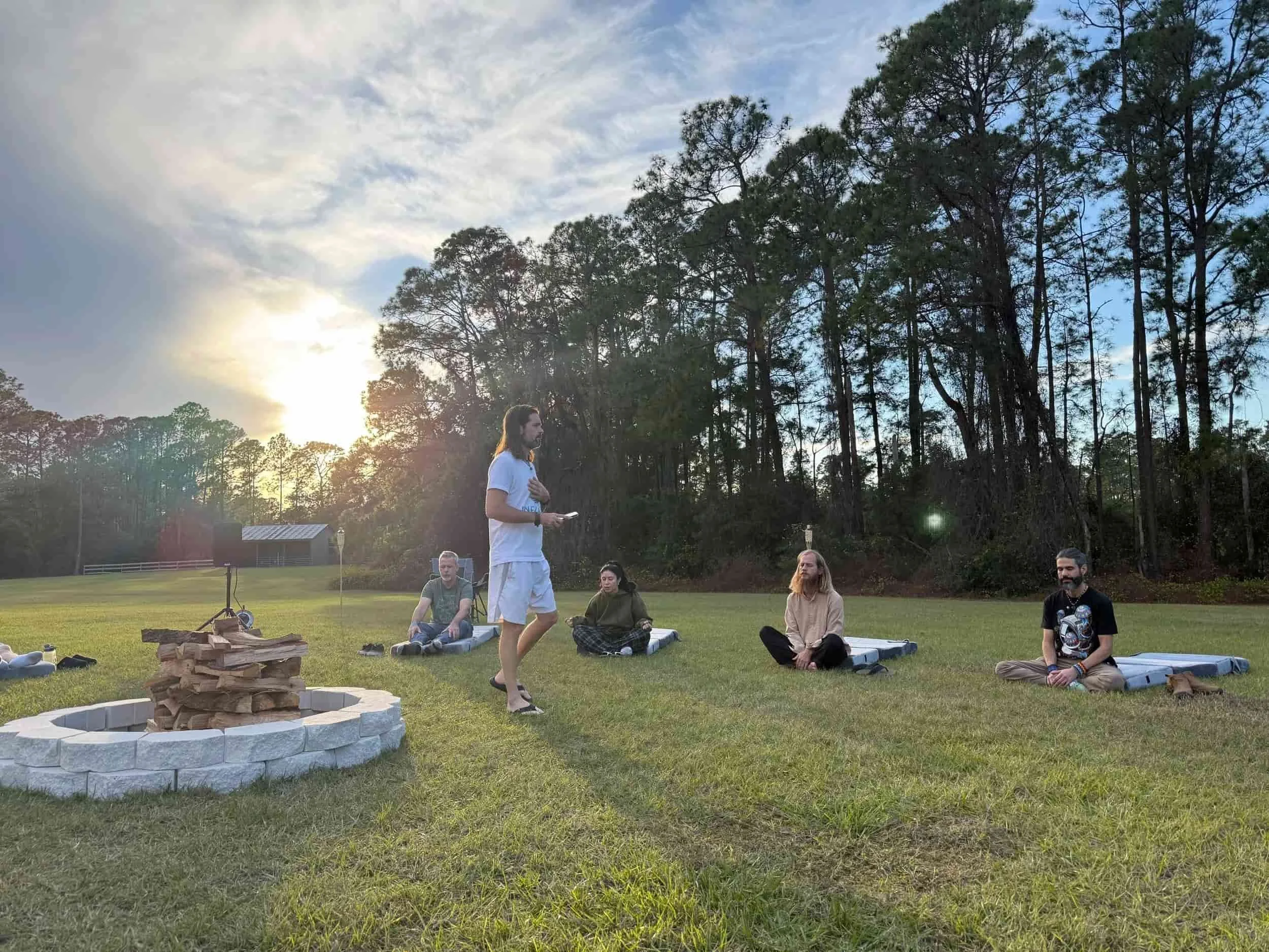 A group of people sitting on the grass outdoors, some with laptops and blankets, with a person standing and speaking. The sun is setting behind trees in the background.