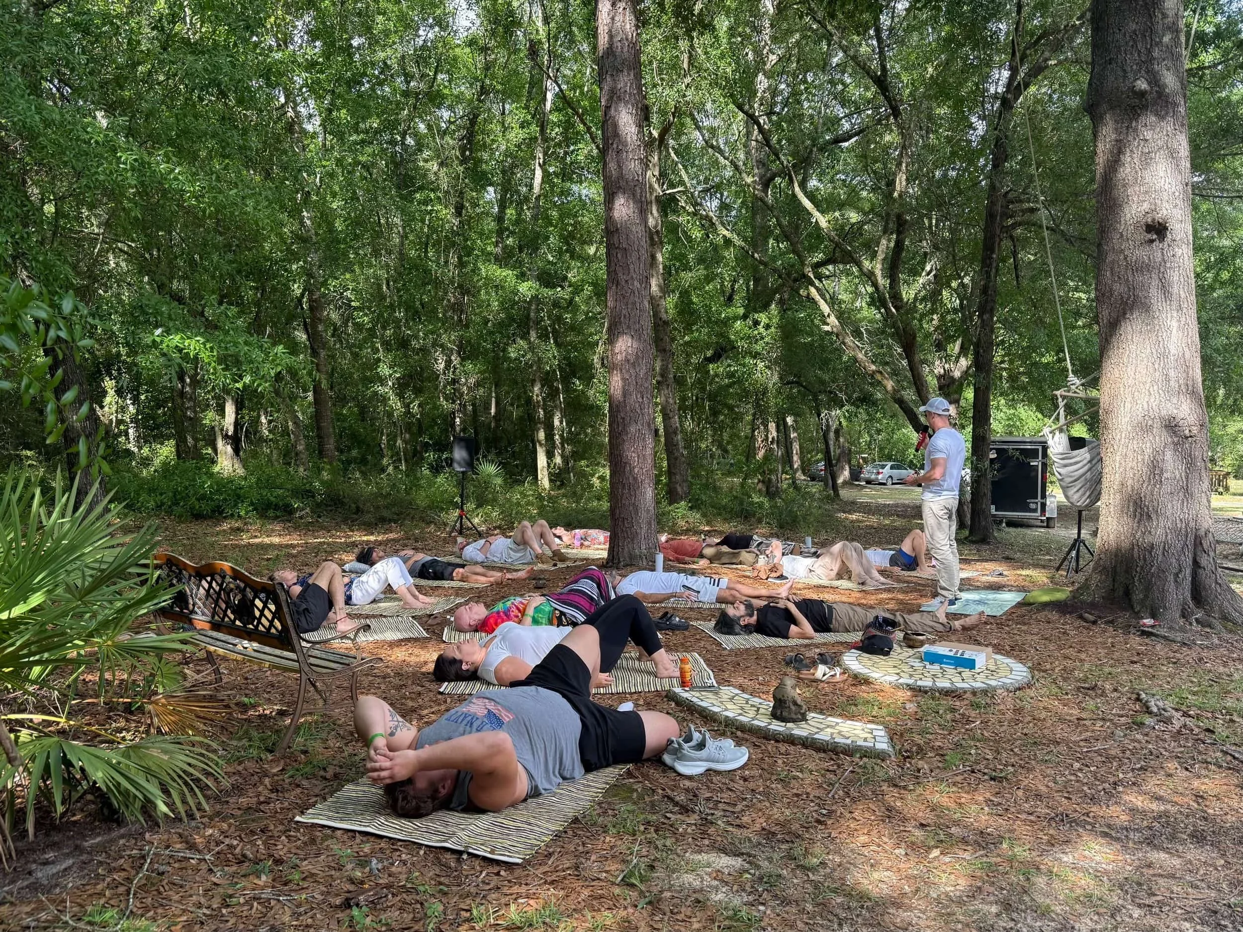 People practicing yoga outdoors in a wooded park area, lying on mats on the ground with trees and parked cars in the background.