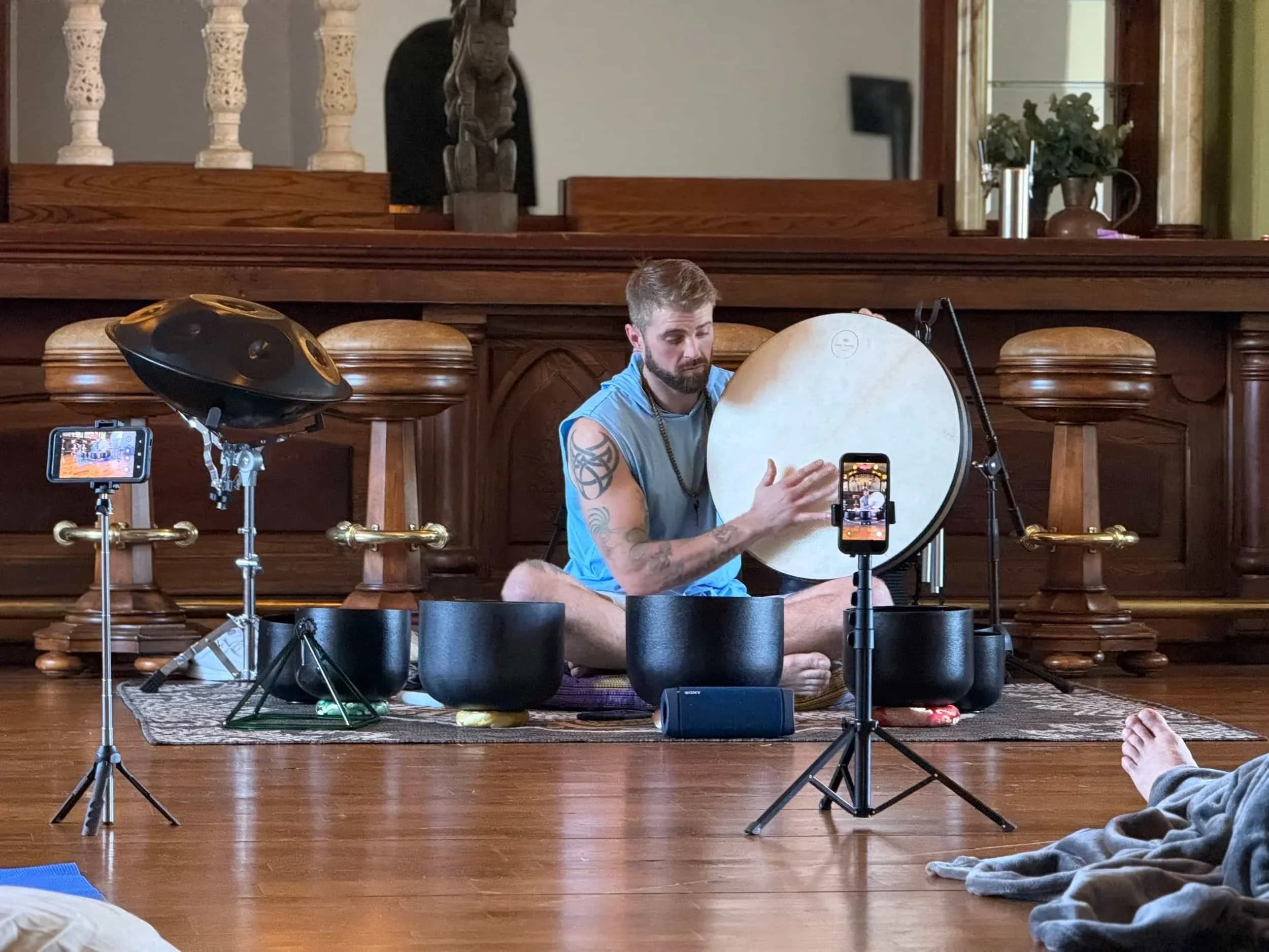 A man sitting cross-legged on a wooden floor playing a gong with a mallet, surrounded by various percussion instruments, in a room with wood furniture, a mirror, and decorative items.