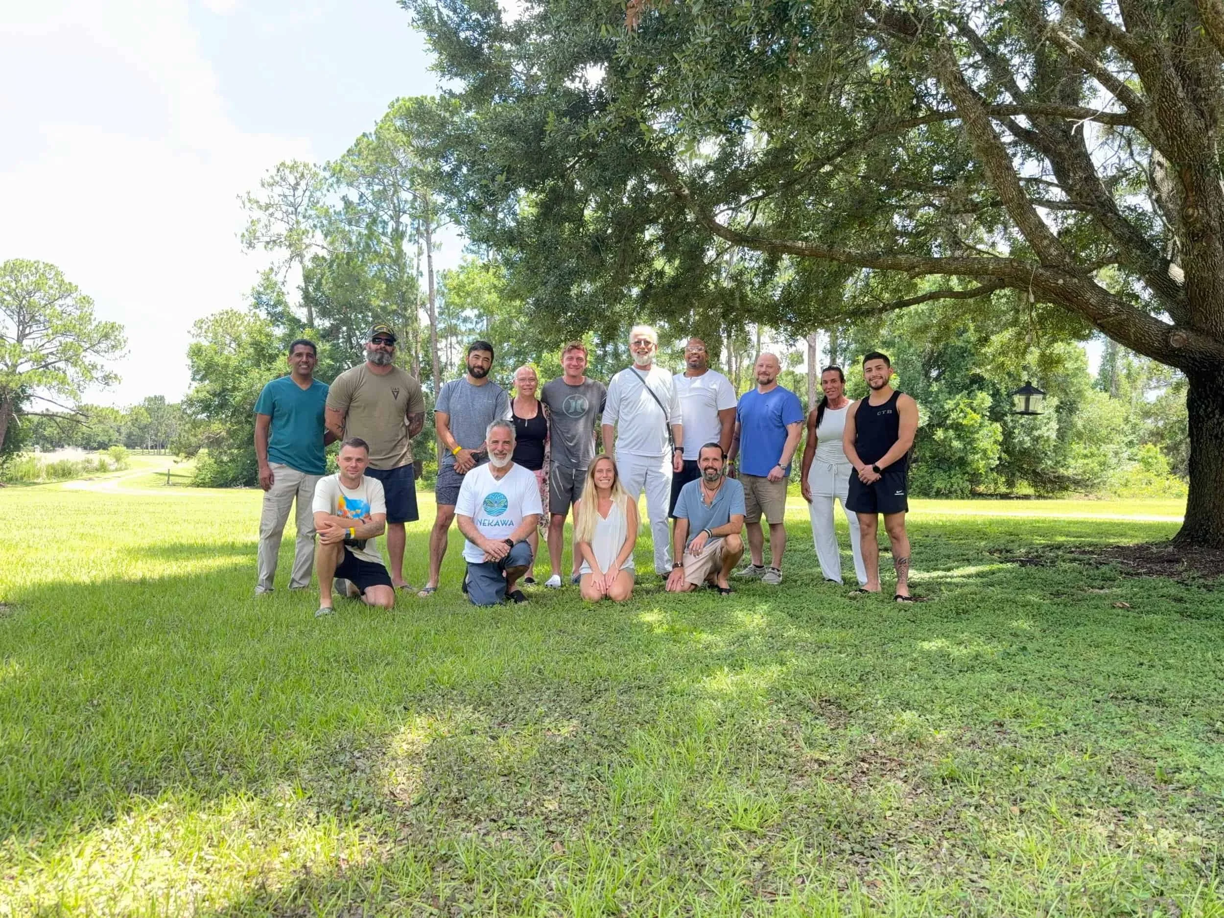 A group of fifteen people, including men and women of various ages, posing outdoors on a grassy area with large trees and a sunny sky in the background.