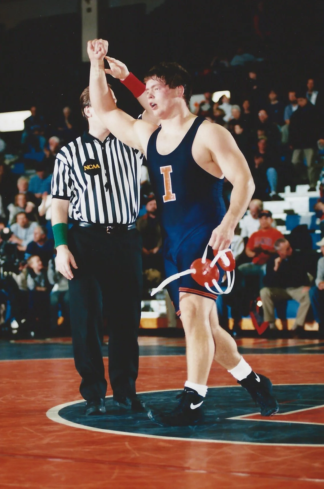 A male swimmer in a blue University of Illinois wrestling singlet being declared winner, with a red towel around his waist and holding a red heart-shaped balloon with white balloons attached, being congratulated by a referee in a black and white striped shirt, in a wrestling arena with spectators in the background.
