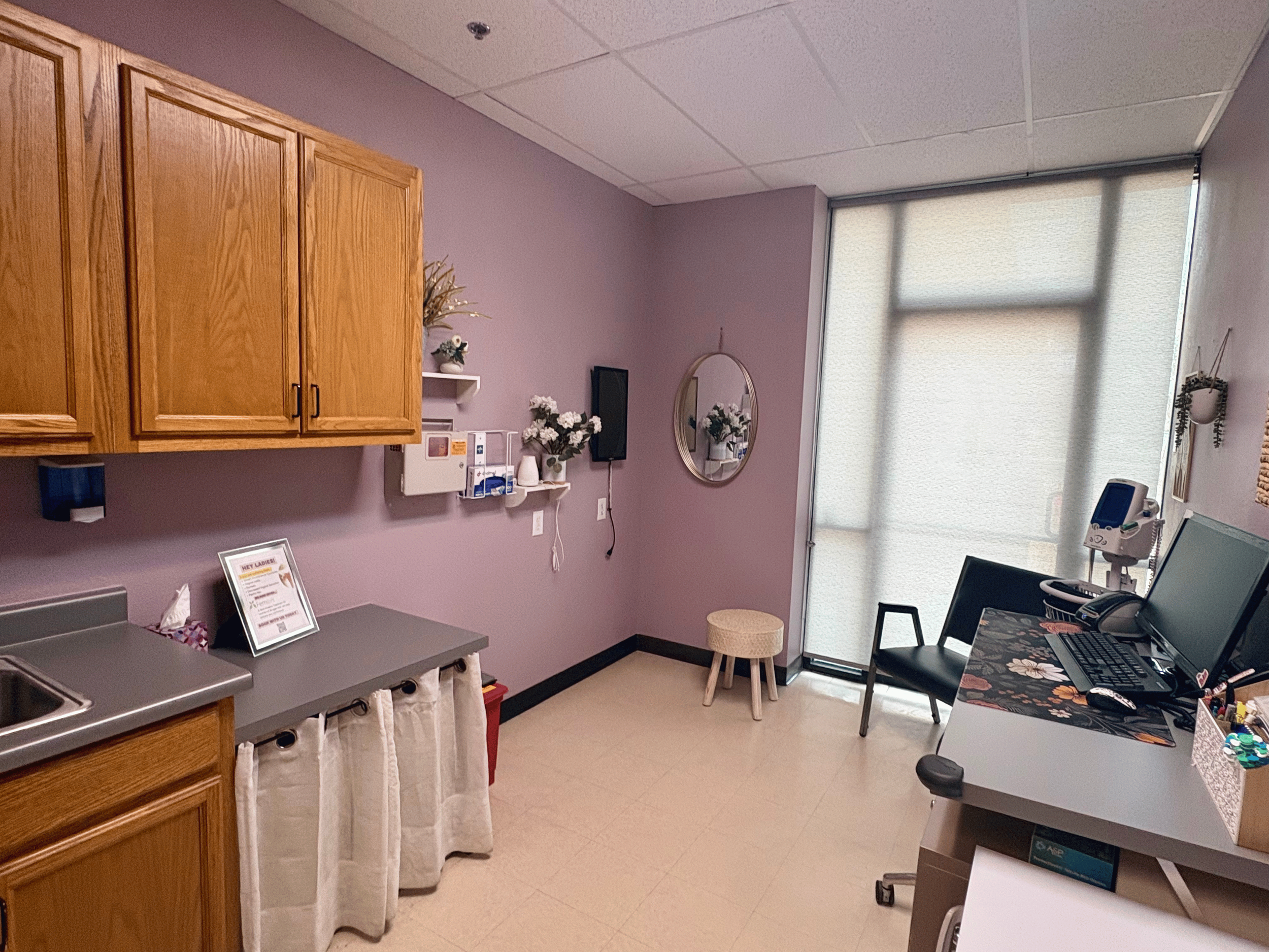 Interior of a medical office or clinic with wooden cabinets, a countertop, and a wall with decorations. There is a window with blinds letting in light. A small stool and chairs are also visible, along with medical equipment and a computer on a desk.