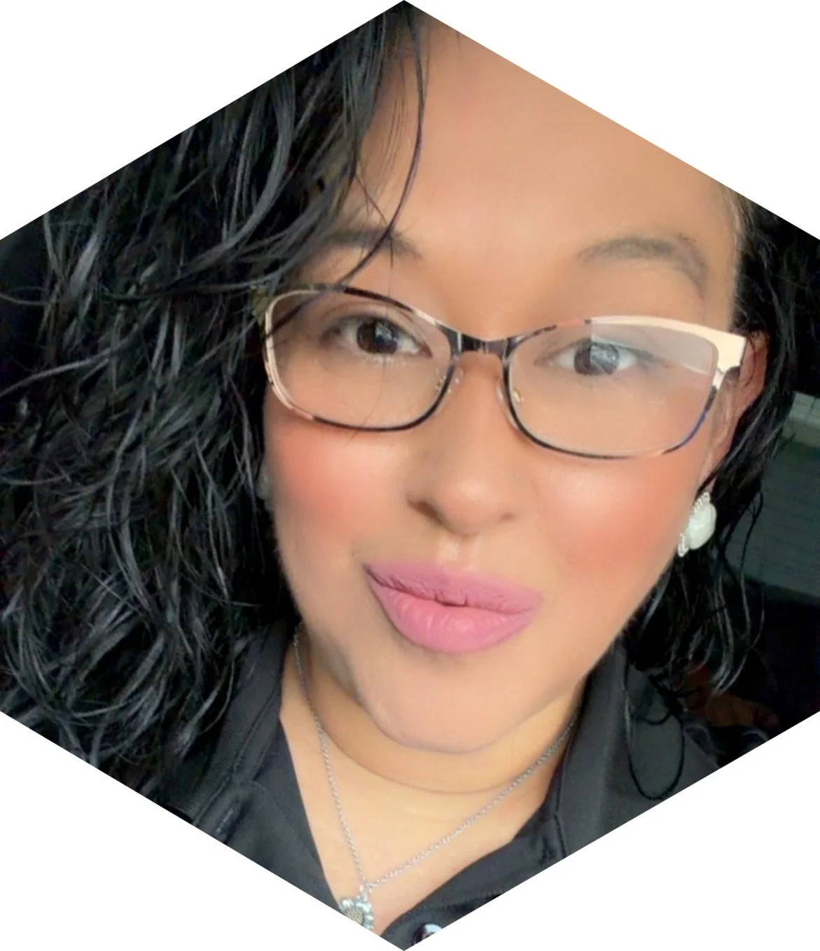 Close-up of a woman wearing glasses, with curly dark hair, pink lipstick, and earrings.