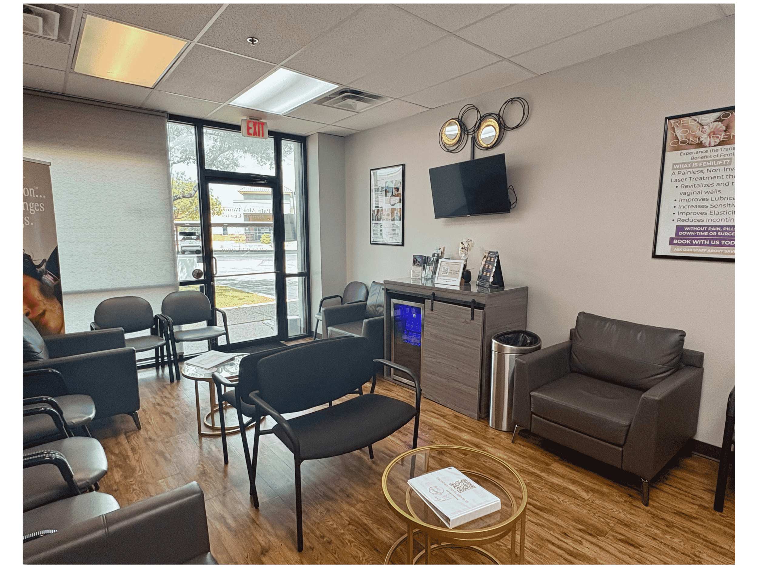 Waiting room with black chairs, a gray armchair, a glass-top side table with books, posters on the walls, a flat-screen TV, and an entrance with glass doors and windows.