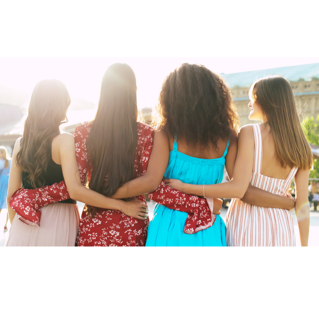 Four women standing close together with their arms around each other, viewed from behind during sunset, outdoor setting.