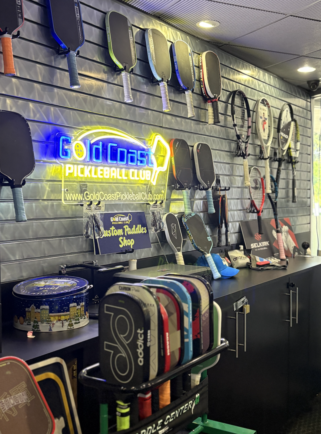 A store display with various pickleball paddles hanging on a wall, a neon sign reading 'Gold Coast Pickleball Club,' and a counter with pickleball equipment and accessories.