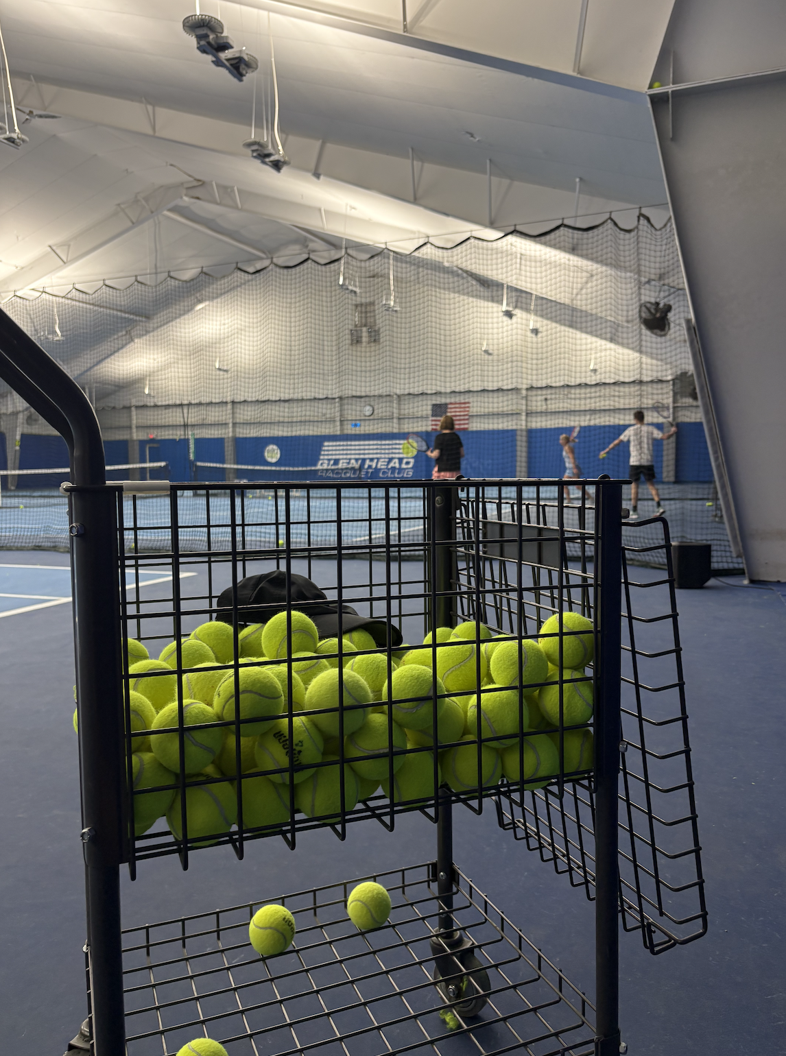 Indoor tennis court with a basket of bright yellow tennis balls in the foreground, and people practicing tennis in the background.