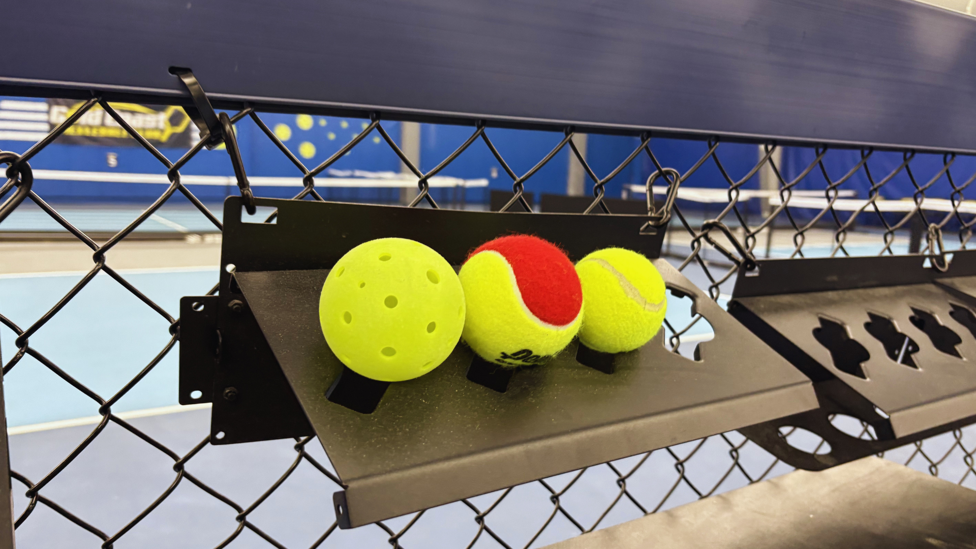 Three tennis balls, one yellow with holes, one red and yellow, and one yellow with a white curved line, resting on a black rack attached to a tennis court fence.