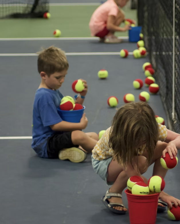 Children sitting on the floor of an indoor tennis court, holding and gathering tennis balls into buckets, with many tennis balls scattered around.