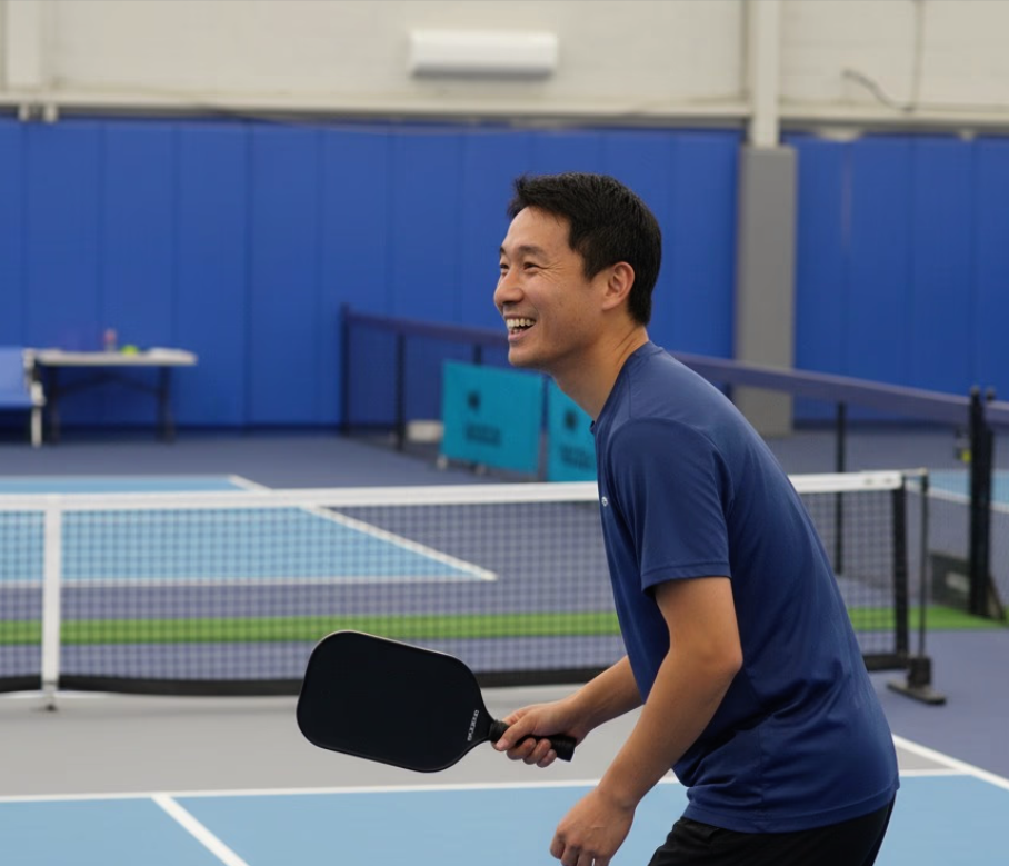 A man with short black hair smiling and holding a ping pong paddle in a recreational indoor ping pong area.