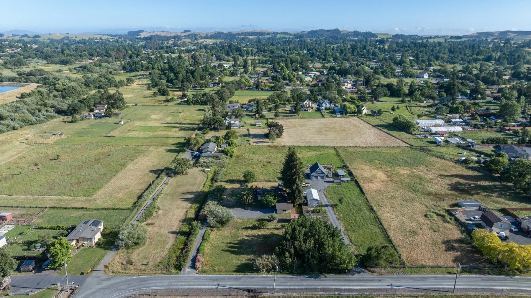 Aerial view of a rural area with scattered houses, large green fields, and trees, with a coastal area in the background under a clear sky.