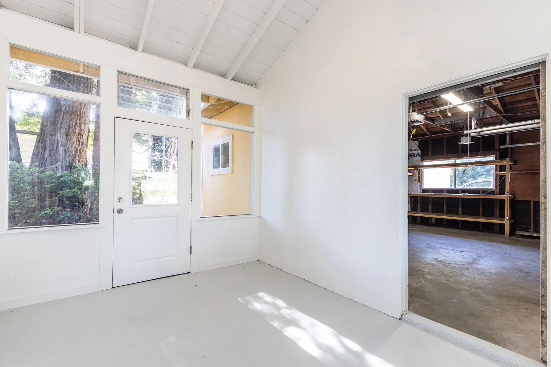 Interior view of a room with white walls and light-colored floor, looking towards an open doorway revealing an unfinished garage with exposed wooden beams and a window.