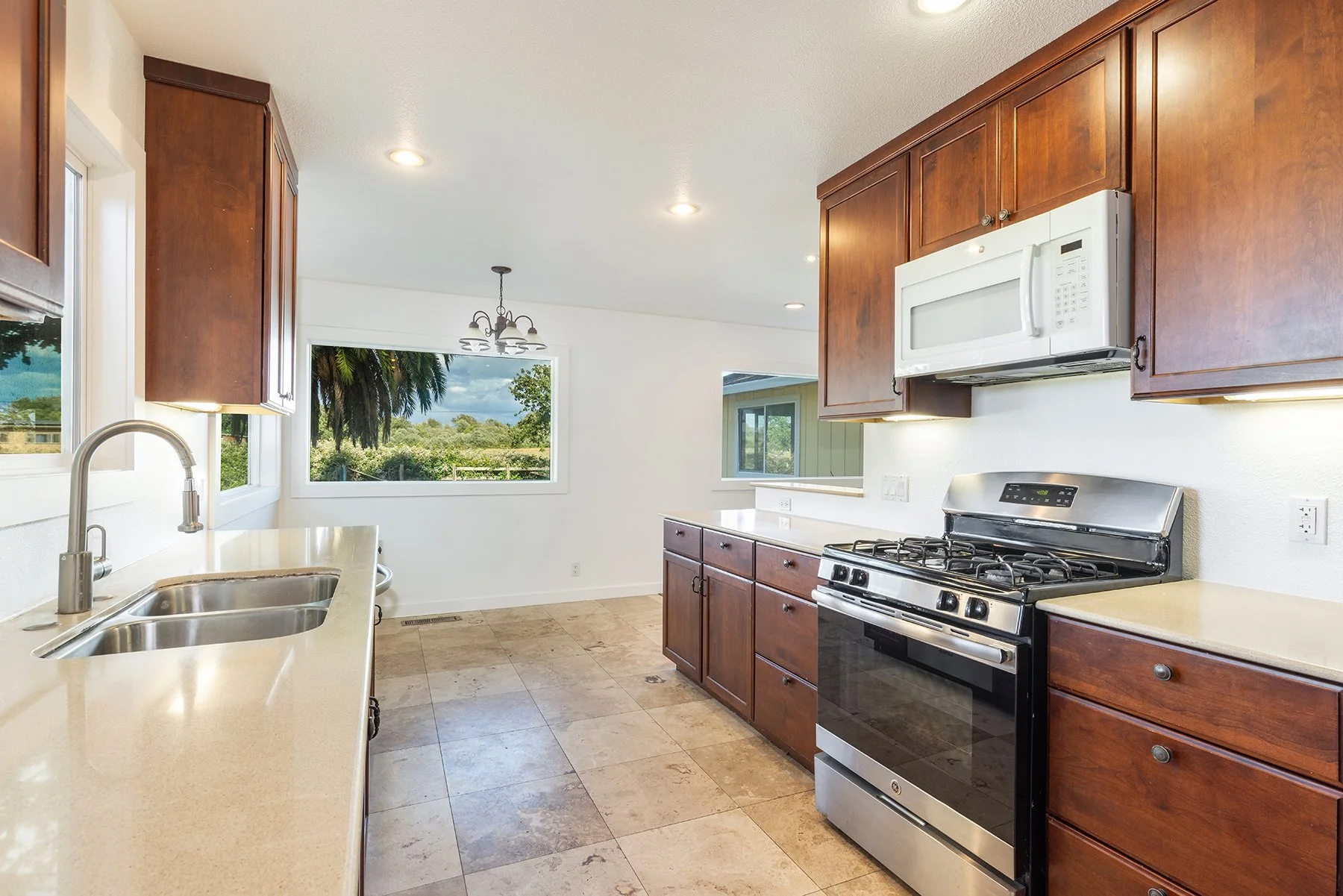 Kitchen with wooden cabinets, stainless steel stove, microwave, beige countertops, double sink, and large window showing trees outside.