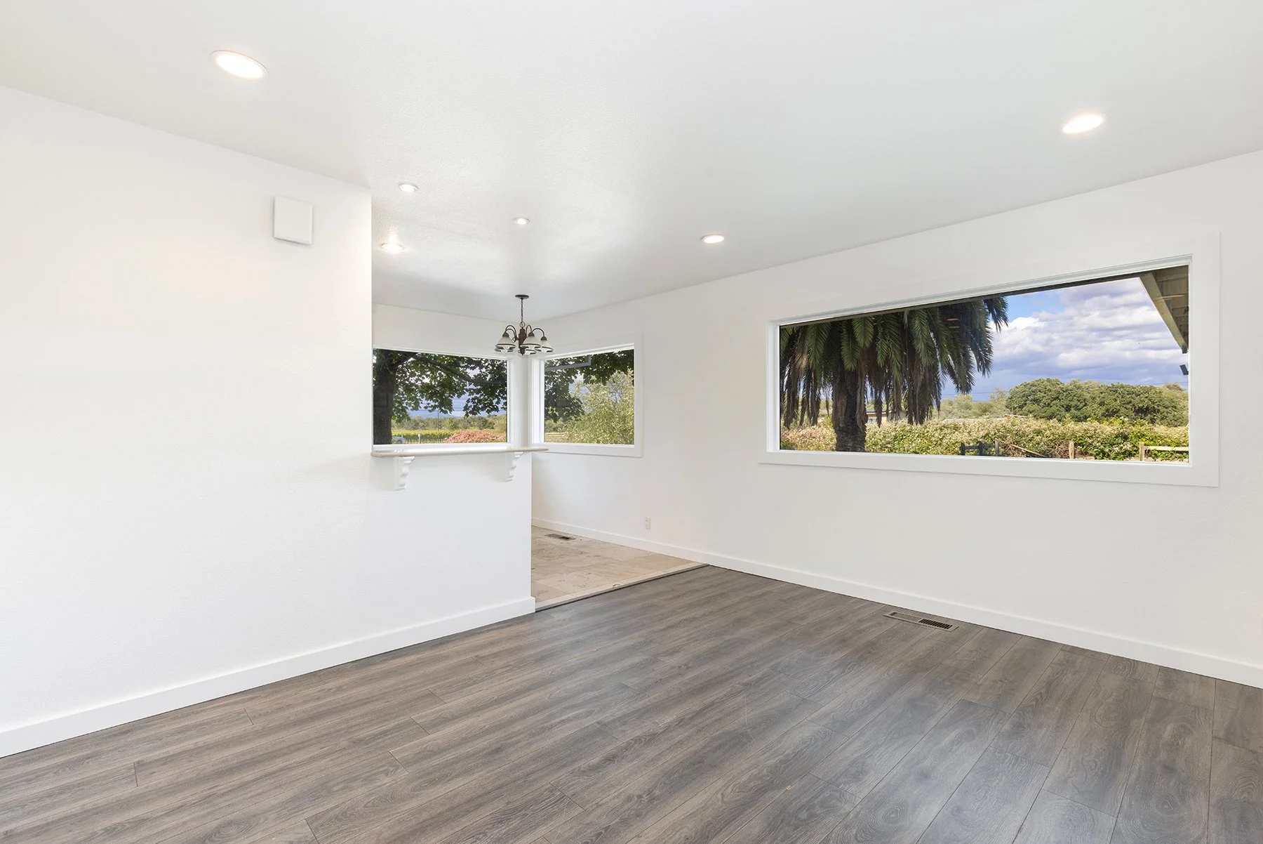 Empty living room with large windows showing outdoor trees and greenery, white walls, and wood flooring.