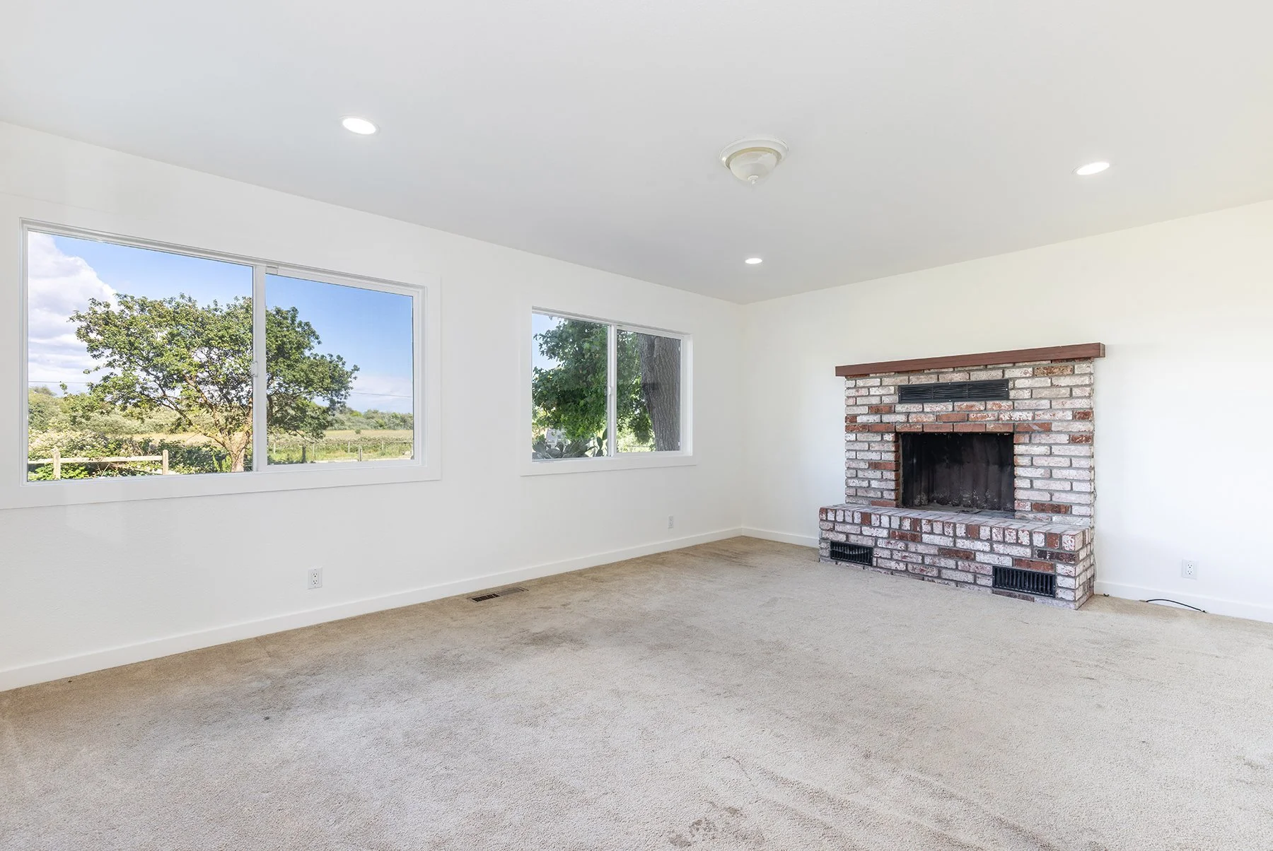 Empty living room with beige carpet, white walls, two windows showing trees outside, and a brick fireplace with a wooden mantle.