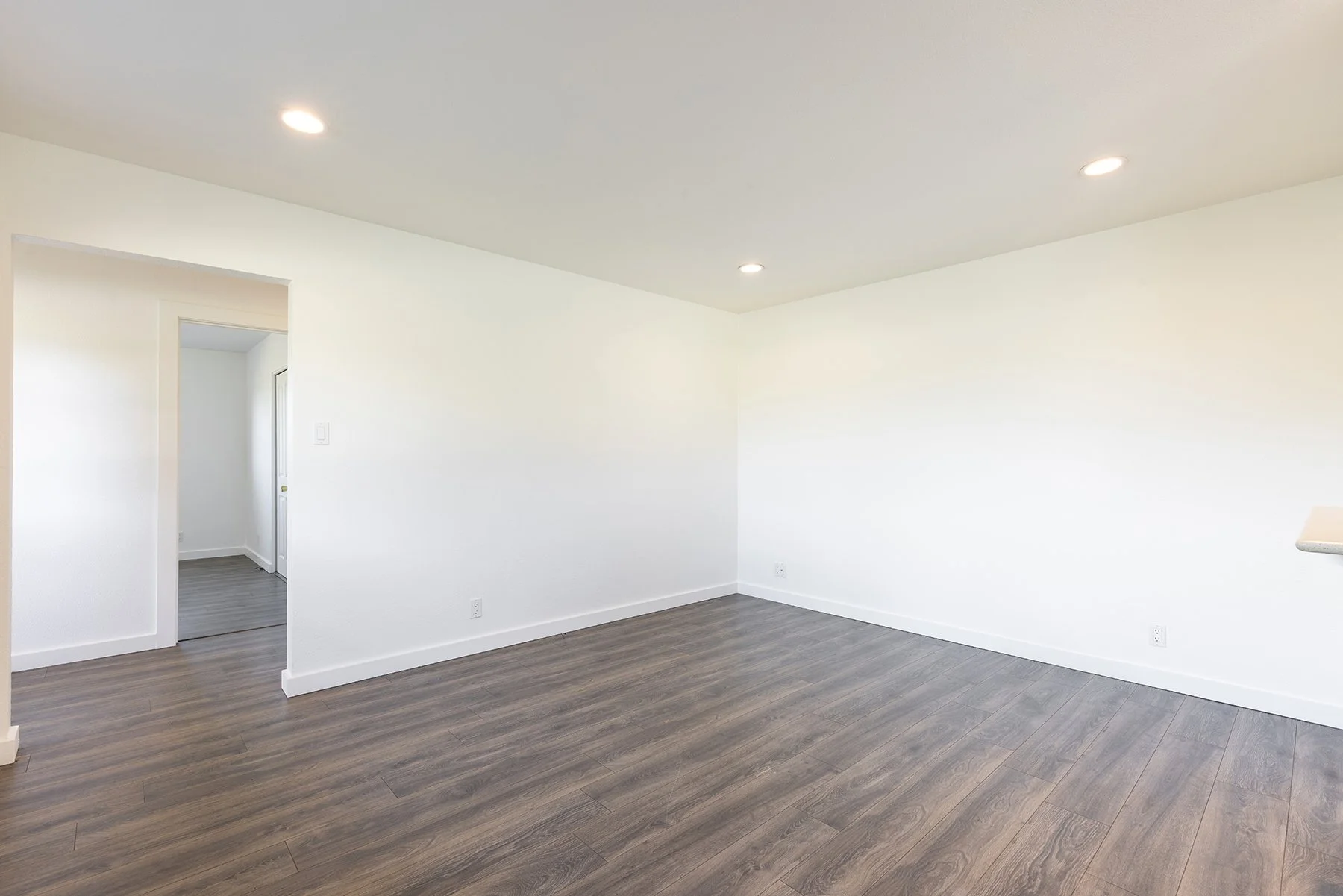 An empty living room with white walls, a white ceiling with four recessed lights, dark wood flooring, and an open doorway leading to a hallway.