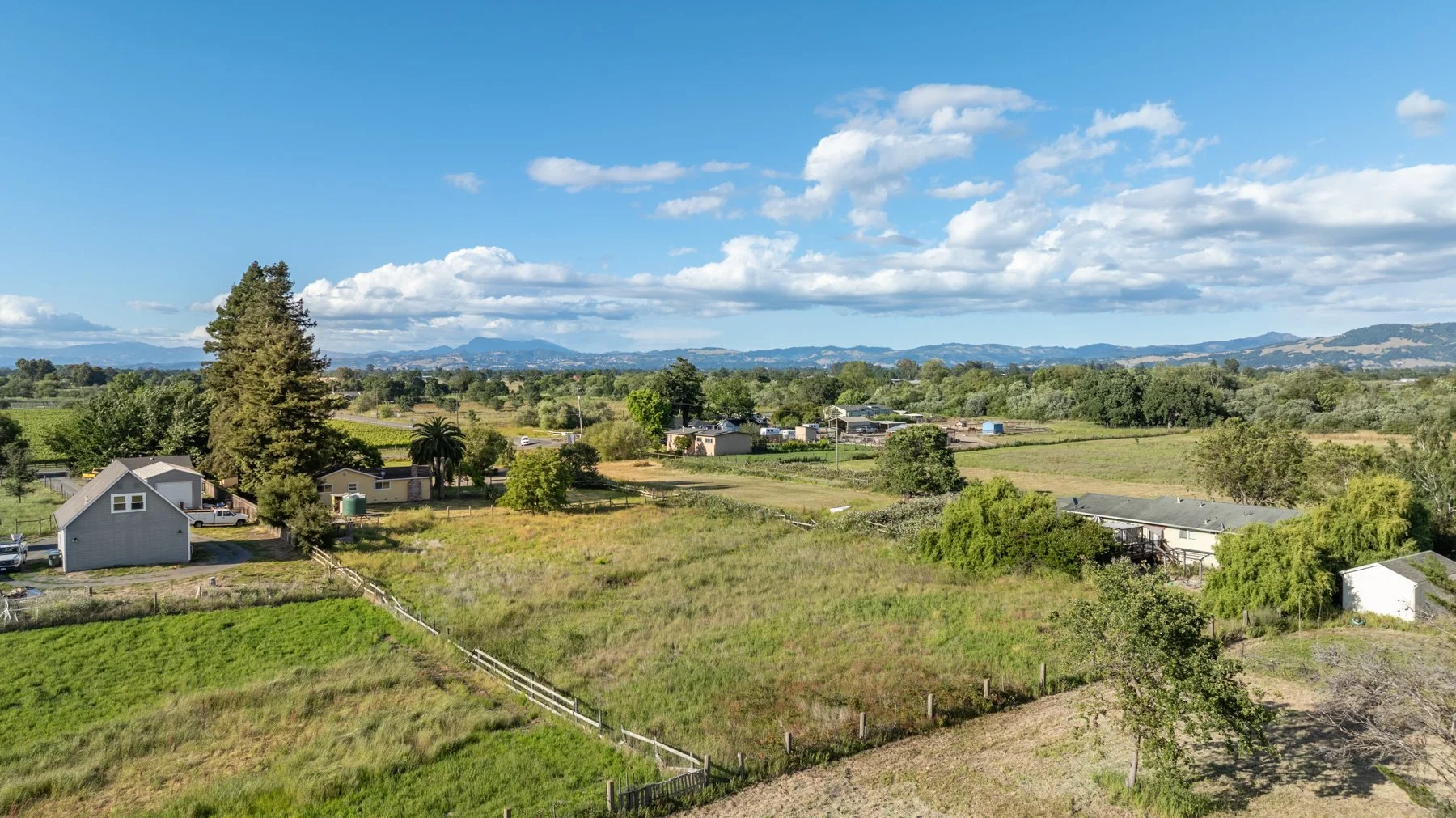 Scenic view of a rural landscape with houses, trees, green fields, and distant mountains under a partly cloudy sky.