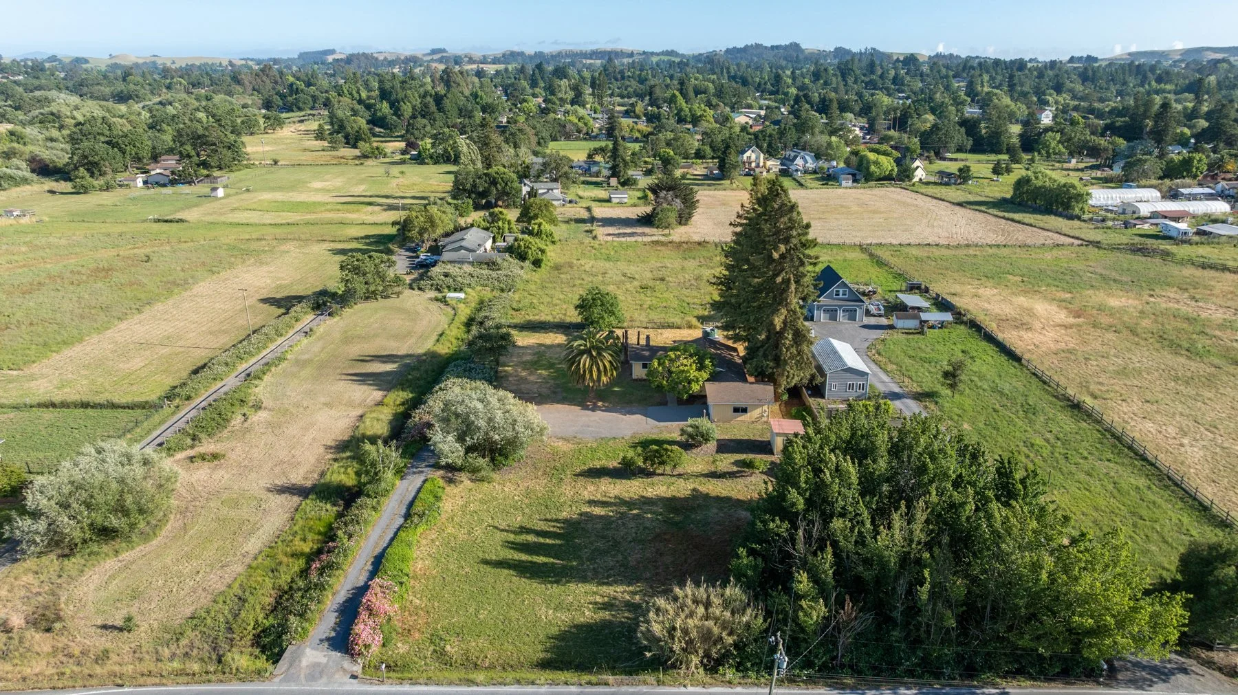 Aerial view of a rural area with farmhouses, open fields, trees, and greenhouses, under a clear sky.