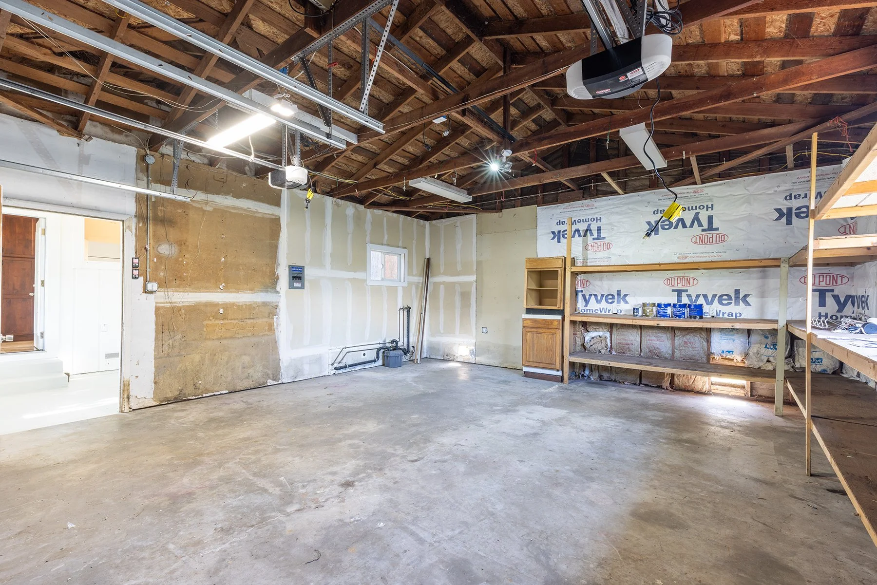 An unfinished garage with exposed wooden beams and insulation, concrete floor, and partially installed electrical and garage door systems. There are shelves with paint cans and construction supplies.
