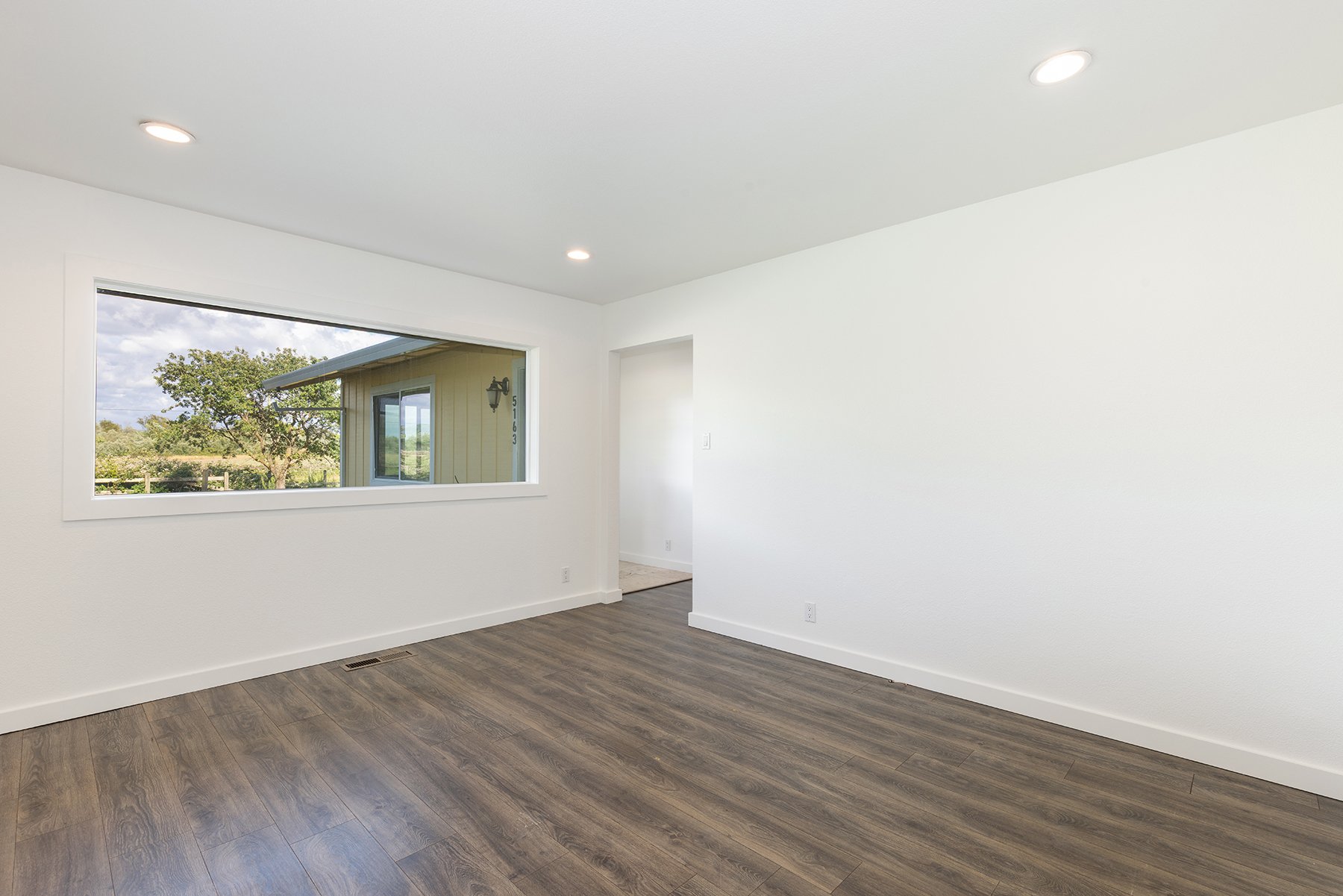 Empty room with white walls, wood flooring, a large window showing a view of a house exterior and trees, and ceiling lights.