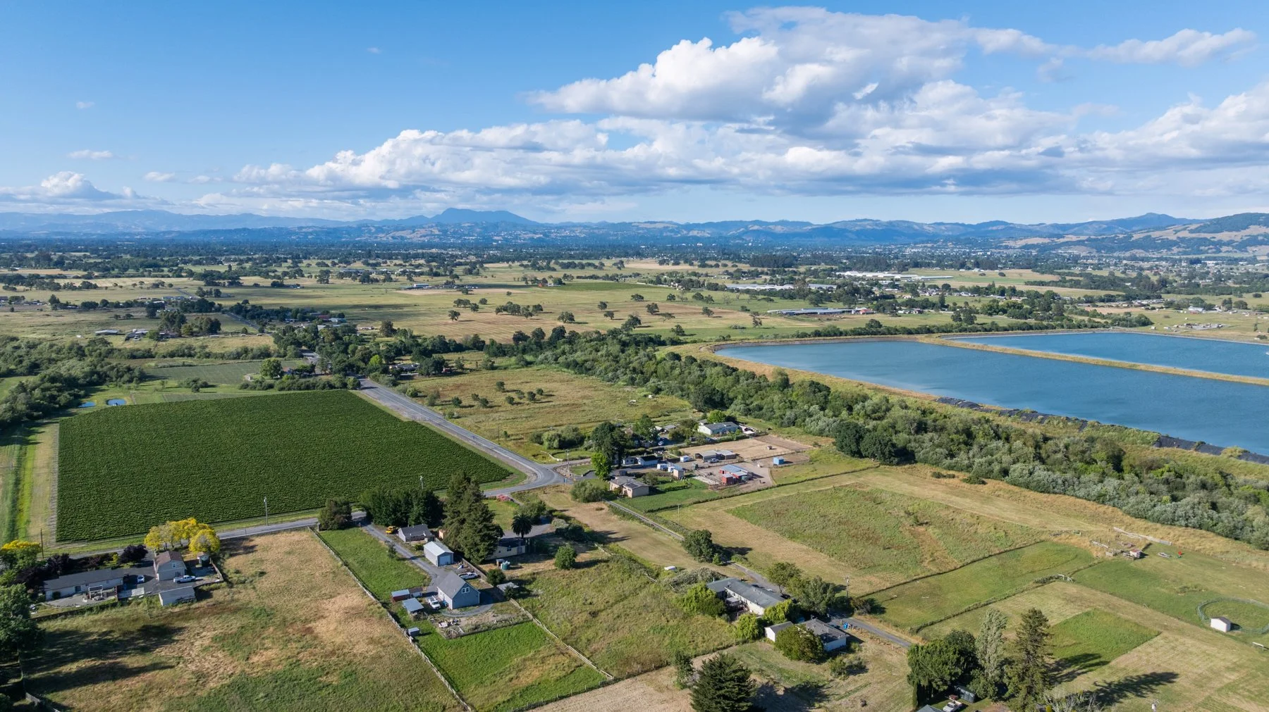 Aerial view of a rural landscape featuring farmland, a vineyard, a large body of water, and distant mountain ranges under a partly cloudy sky.