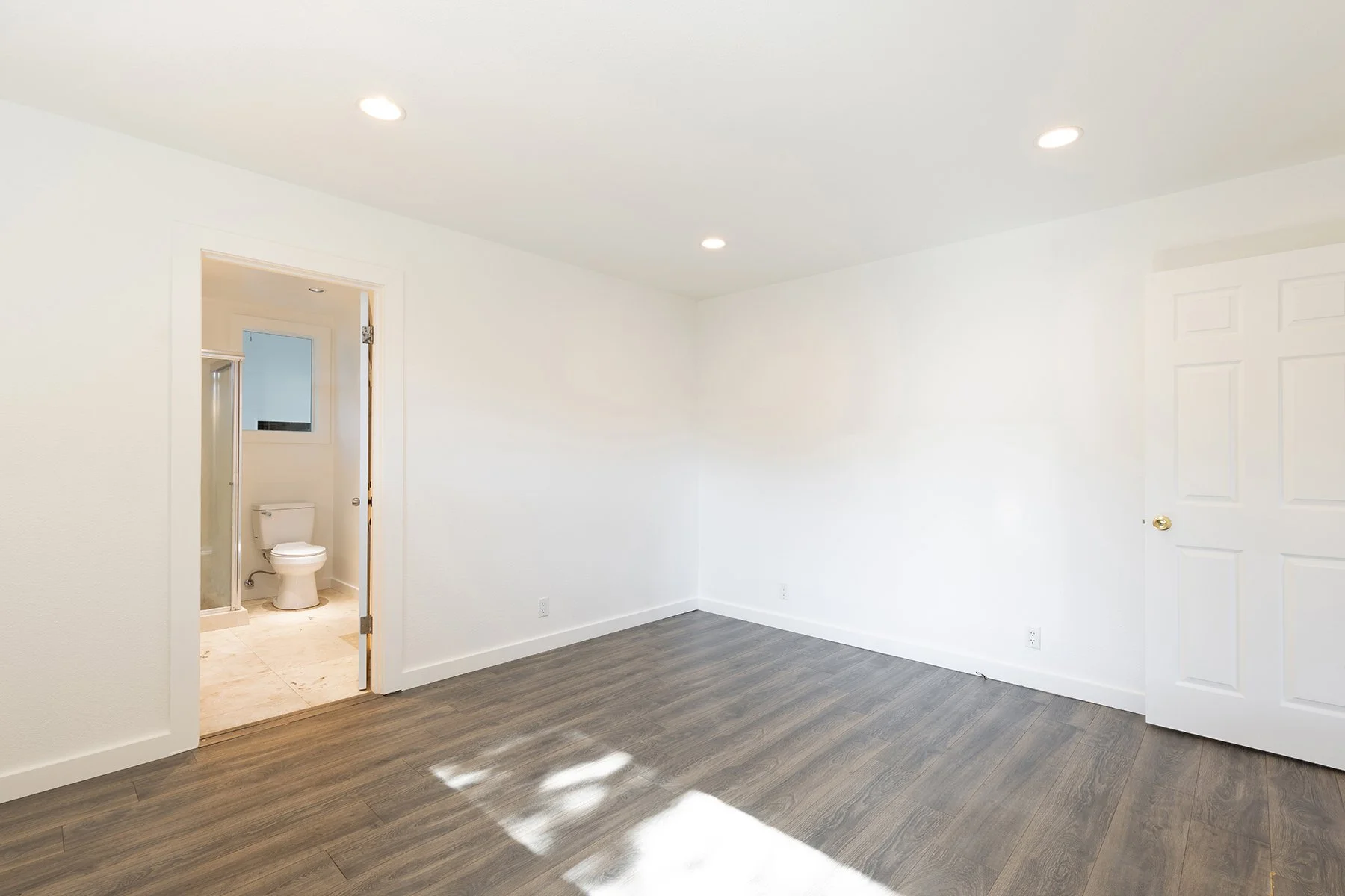 Empty room with white walls, wood flooring, a door to a bathroom with a toilet, and a closet door.