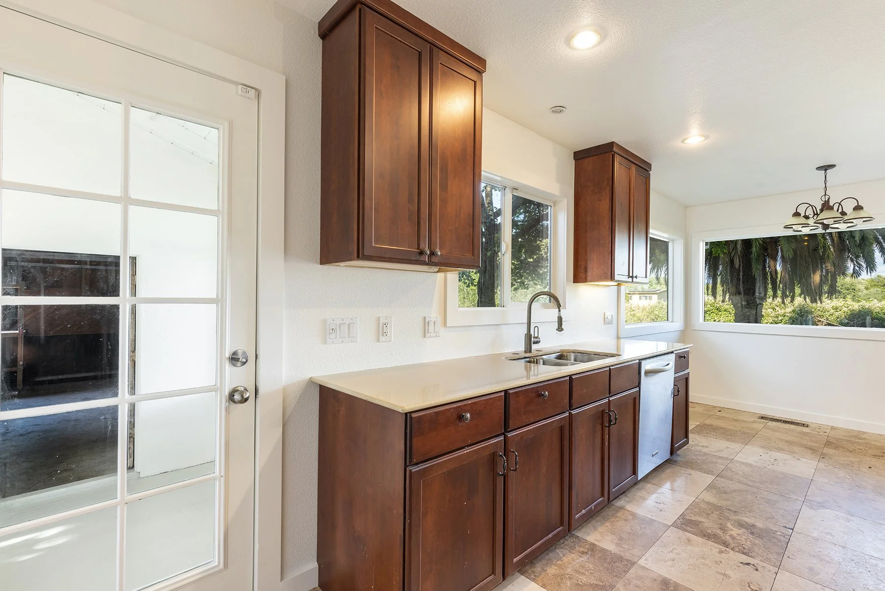 Kitchen with dark wood cabinets, cream countertops, and a double sink under windows. There is a white door with glass panes, a chandelier, and a view of trees outside.