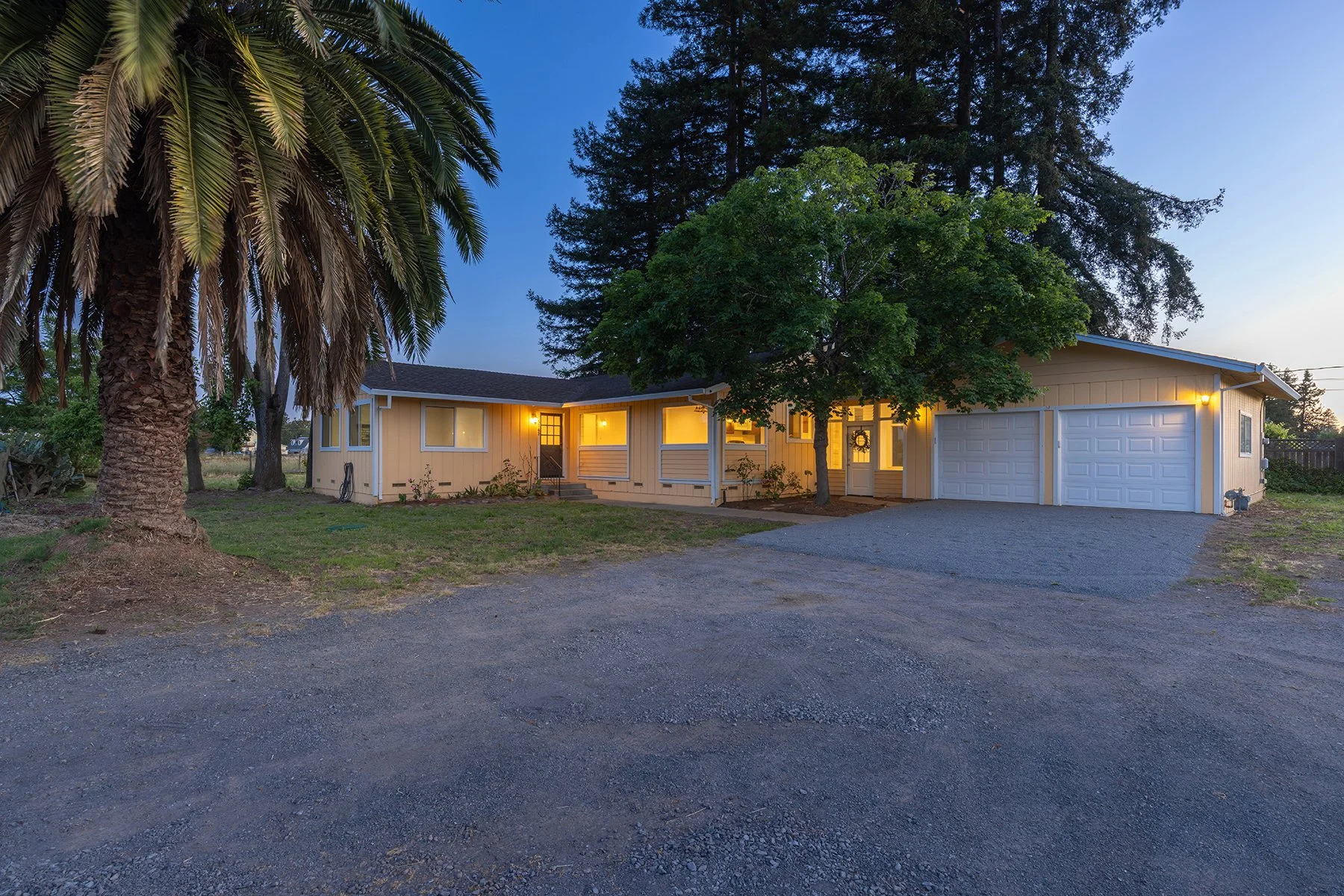 A yellow house with a garage in a yard with trees and a gravel driveway during dusk, with warm interior lights shining through the windows.