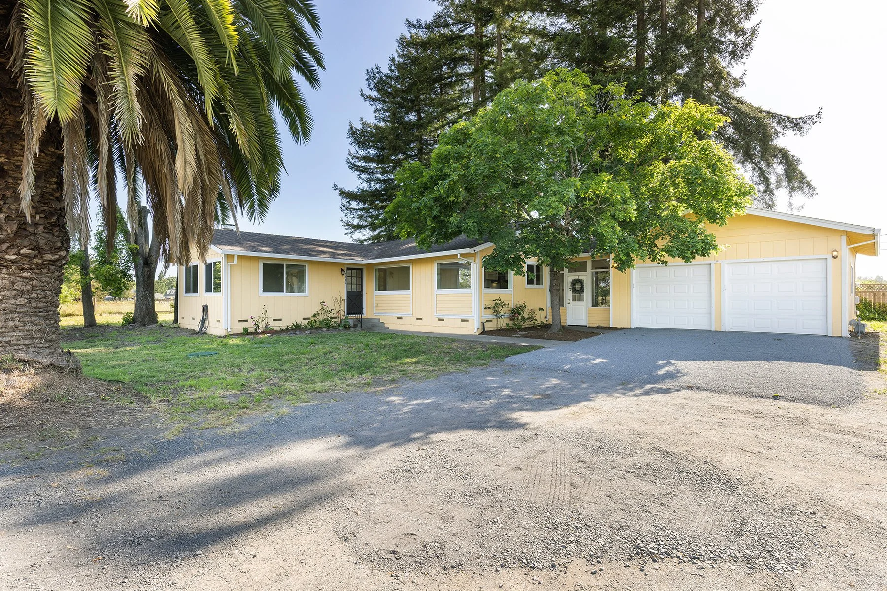 A yellow house with white trim and a two-car garage, surrounded by trees and situated on a gravel driveway.