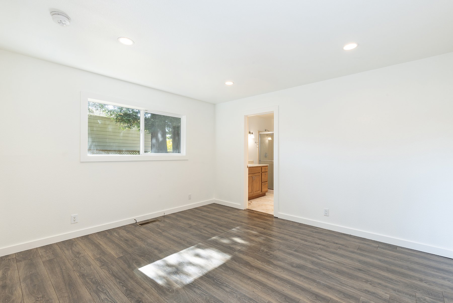 Empty room with white walls, large window, and wood-look flooring, connected to a bathroom with wooden vanity and mirror.