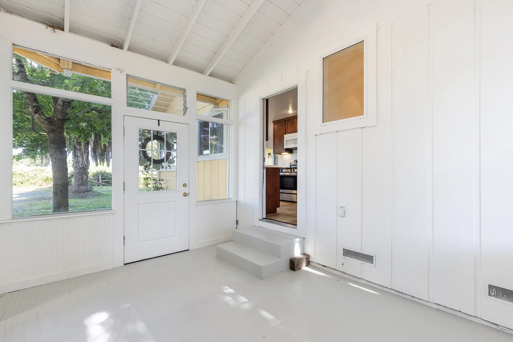 Bright sunlit porch with white walls, windows, and door, leading to a kitchen area with dark wooden cabinets and a stove. Outside view of green trees.