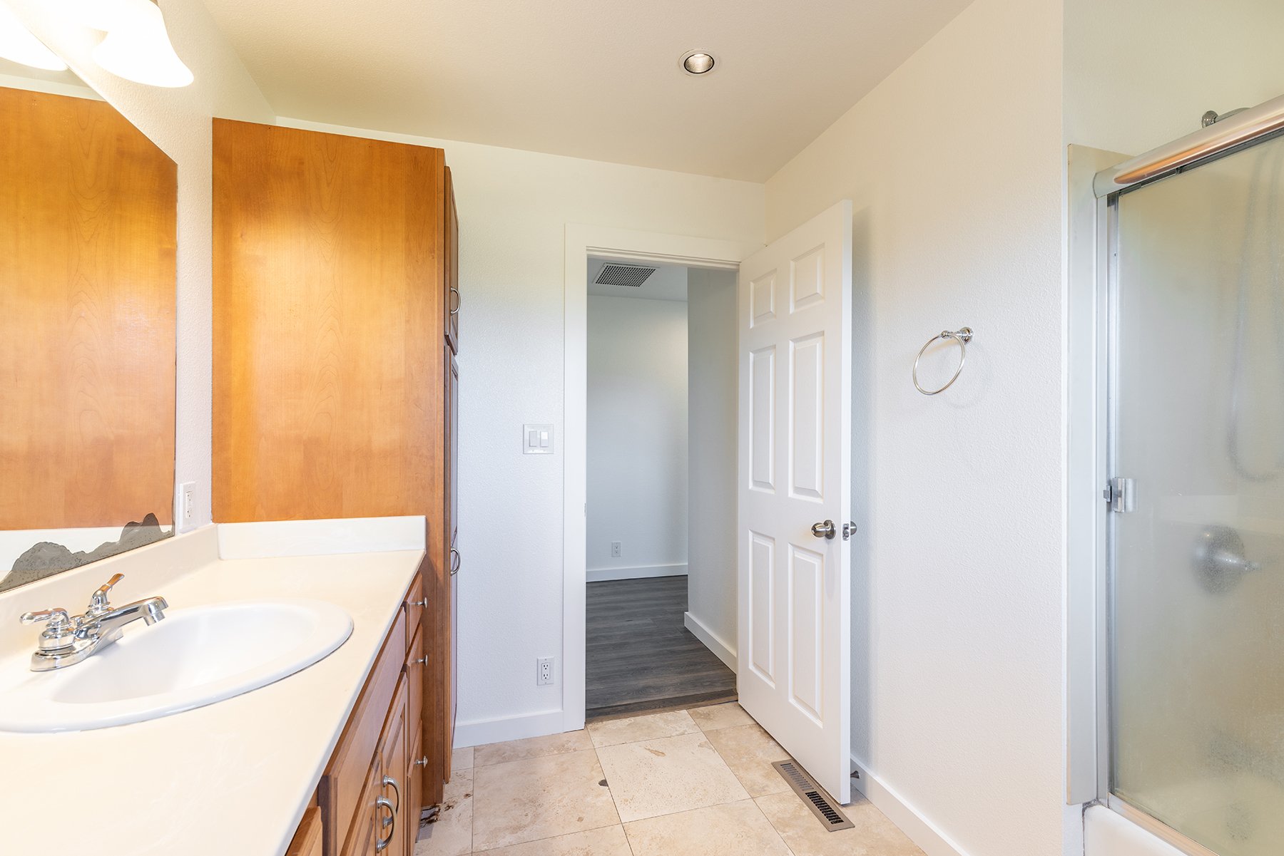 A bathroom with a white countertop and wooden cabinets, a mirror, a white door, a towel ring, and a glass shower enclosure.