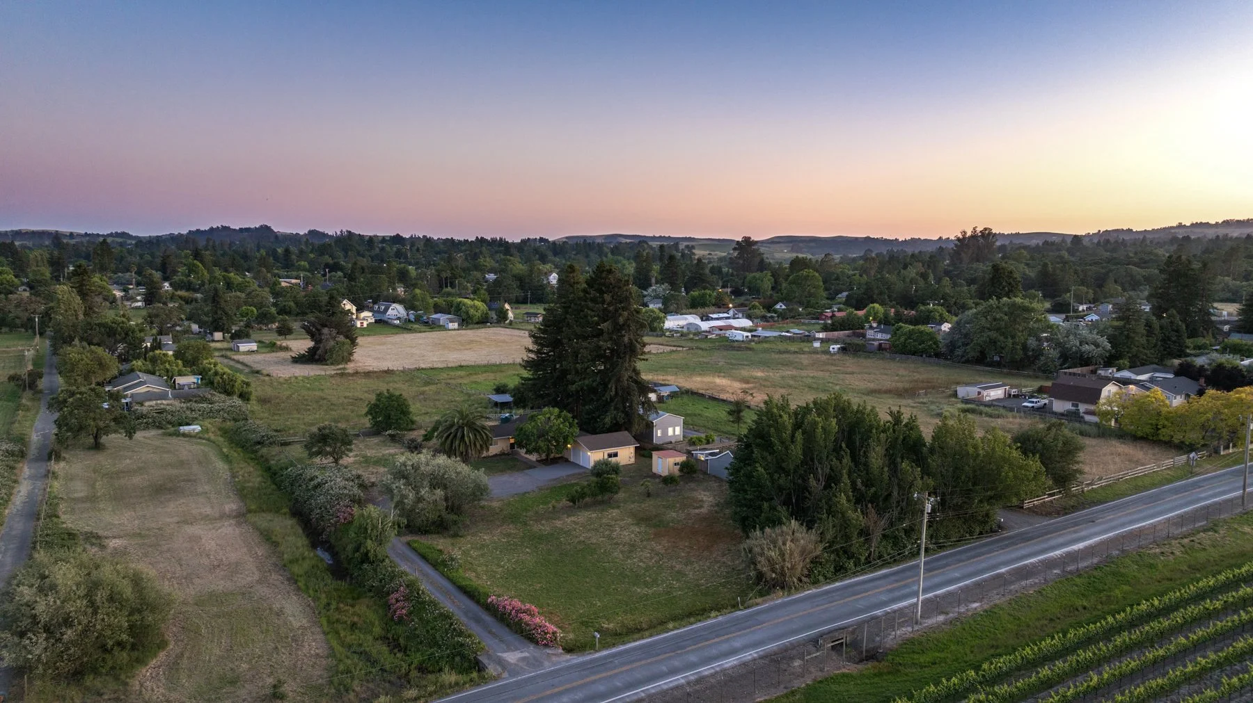 A scenic view of a rural neighborhood at sunset with houses, trees, open fields, and a mountain range in the background.