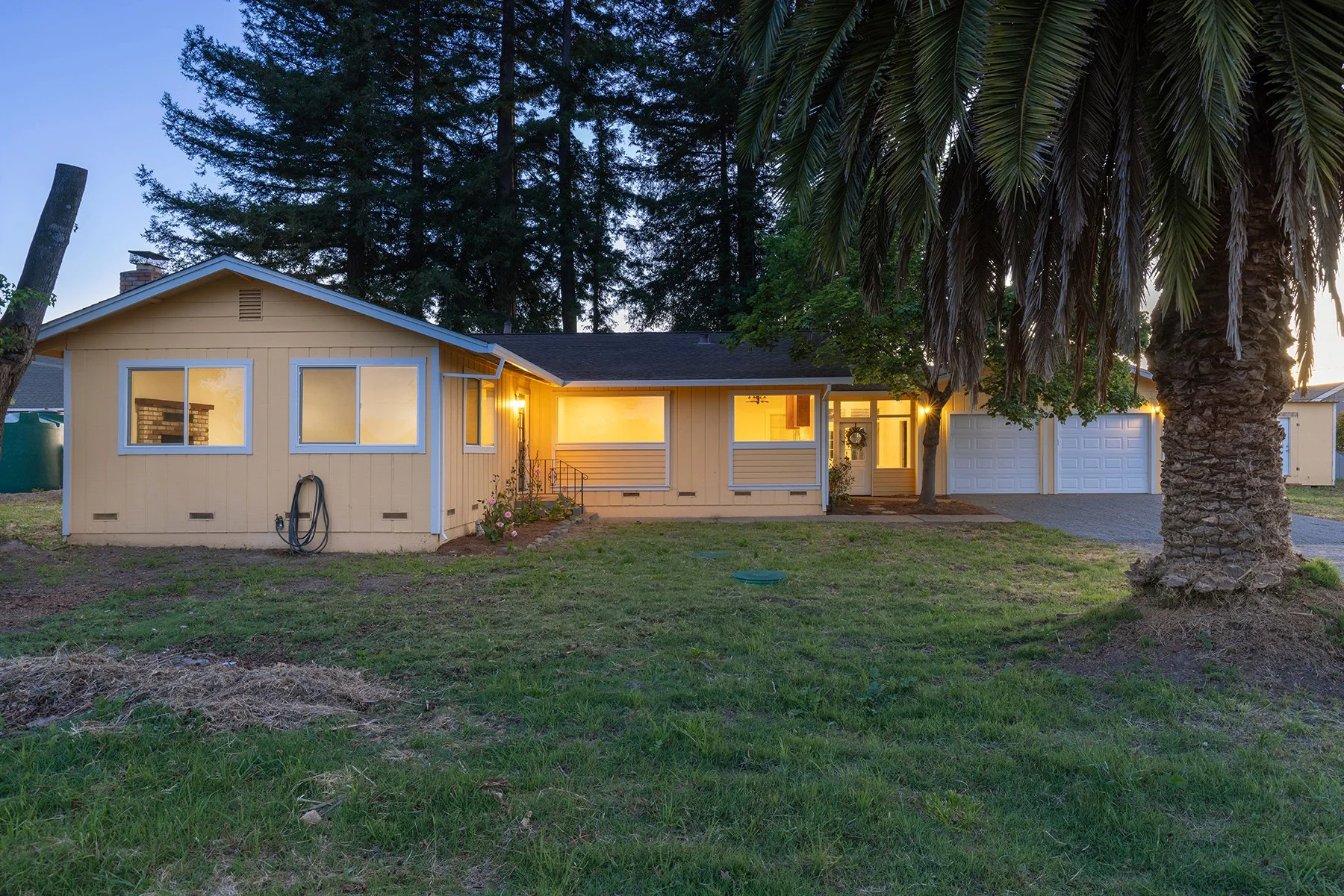 Single-story yellow house with a large palm tree in the front yard, illuminated windows, and a garage, during dusk.