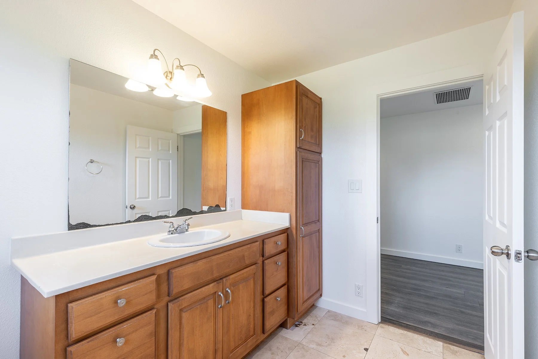 Bathroom vanity with a mirror, wooden cabinets, a white countertop, and a sink, with a door leading to an empty room and a ceiling light fixture.