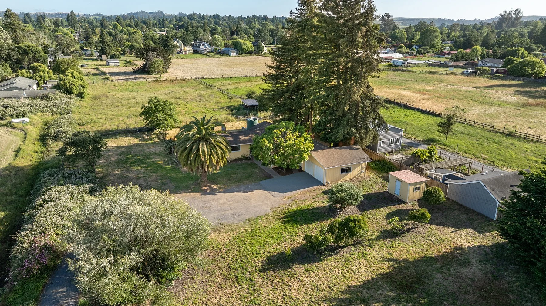 Aerial view of a rural property with a main house, smaller outbuildings, large trees, and open green fields surrounded by a wooden fence, with other houses and fields in the background.