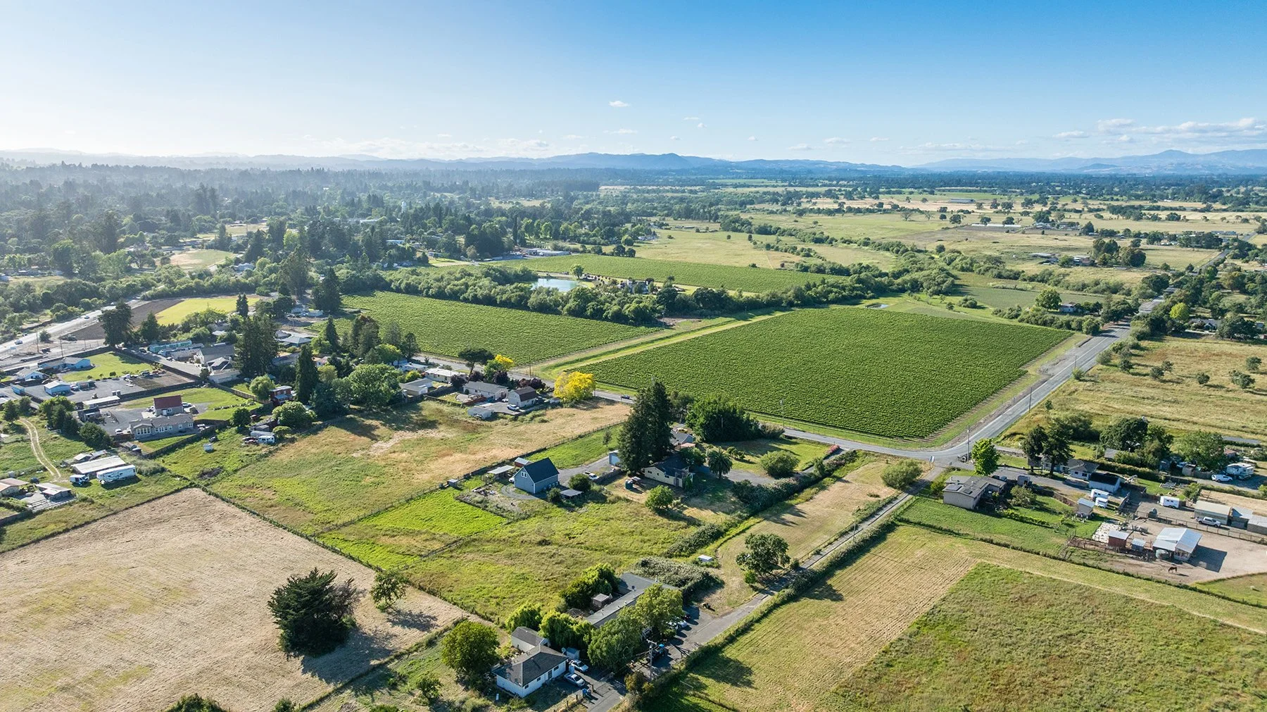 Aerial view of a rural landscape with farmland, houses, roads, and green fields under a clear blue sky.