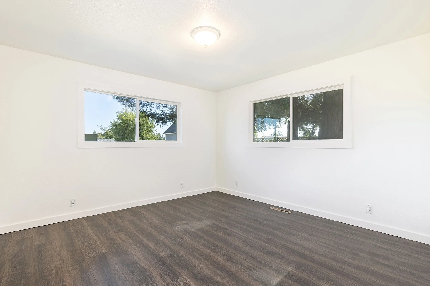 Empty room with white walls, two windows showing trees outside, brown hardwood flooring, and ceiling light fixture.