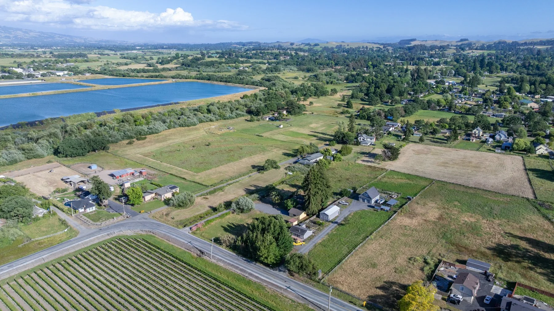 Aerial view of a rural area with houses, farms, and a large body of water, surrounded by lush greenery and fields.
