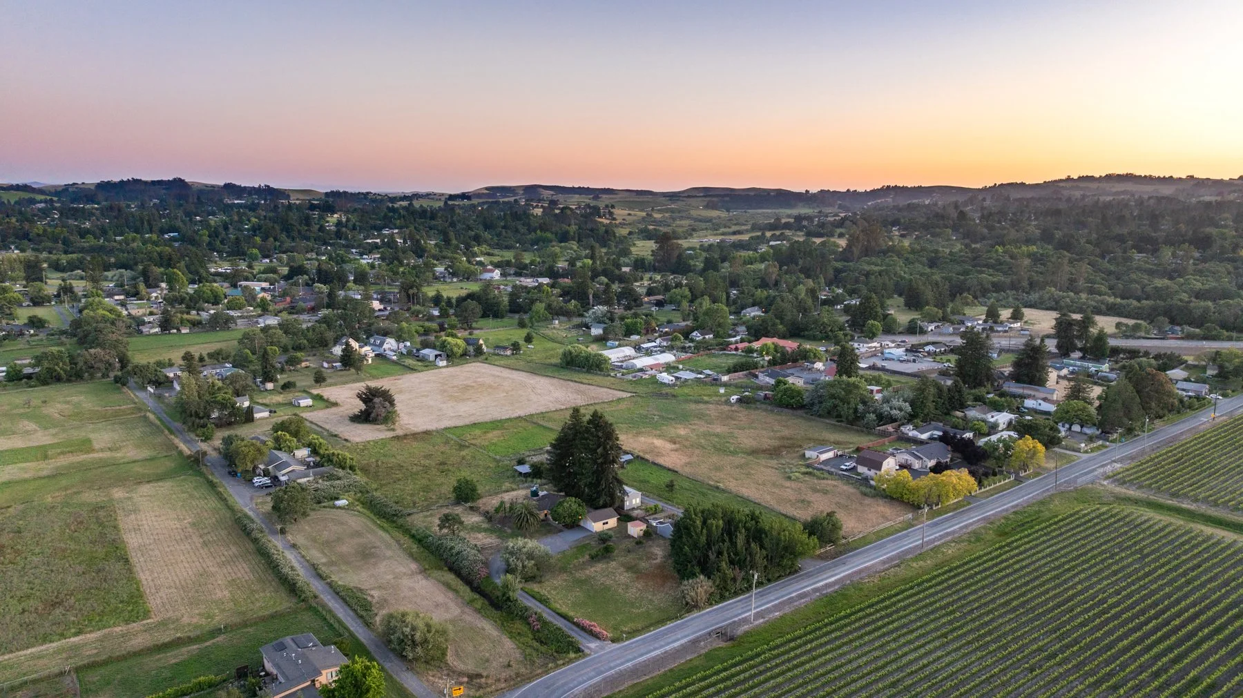 Aerial view of a rural area at sunset with farmland, residential homes, and rolling hills in the background.