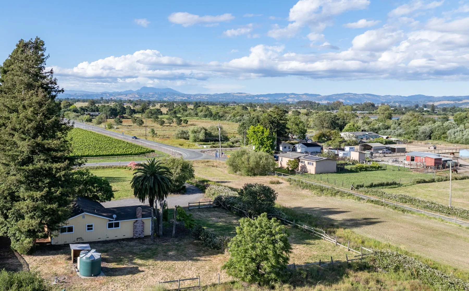 Scenic rural landscape with houses, farm fields, and trees under a blue sky with clouds, hills in the distance.