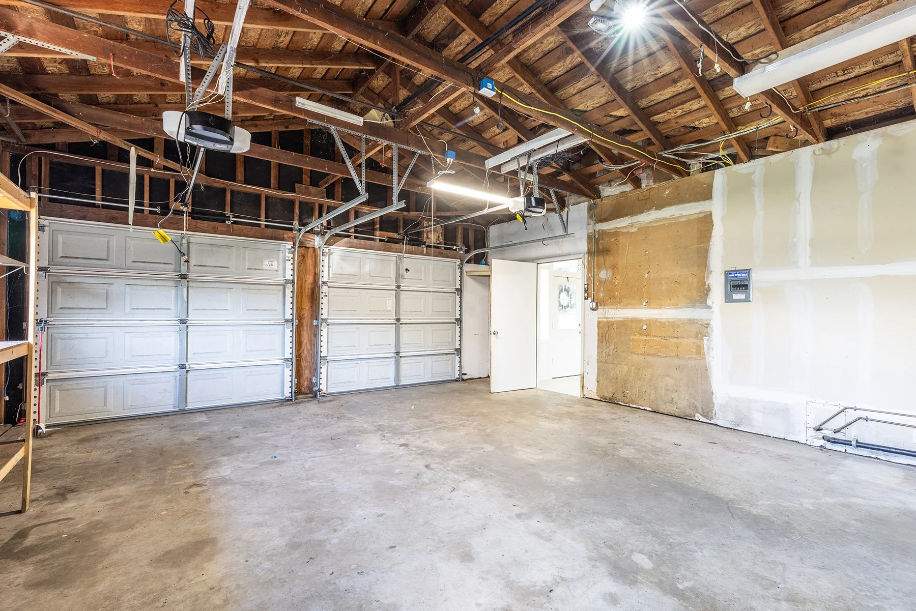 Empty garage with unfinished walls, concrete floor, and a large motorized garage door. The ceiling shows exposed wooden beams and electrical wiring. There is a small open door leading to another room.