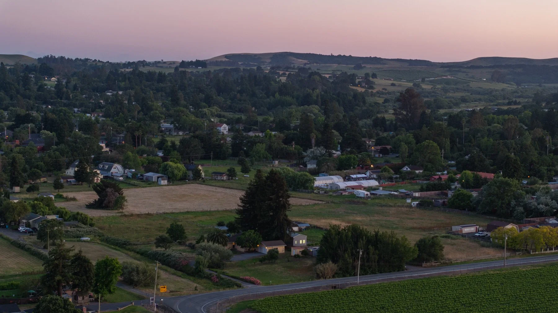 Scenic view of a rural landscape at dusk with rolling hills, scattered trees, houses, farmland, and a winding road in a peaceful countryside setting.