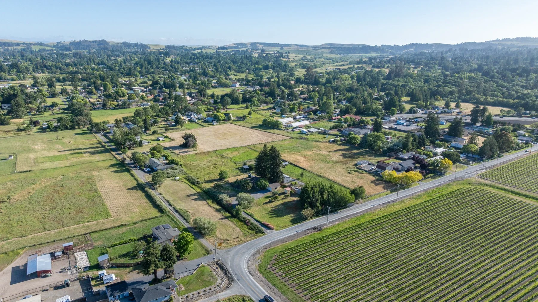 Aerial view of a suburban area with residential houses, green fields, and farmland with rows of crops, surrounded by trees and hills in the background.