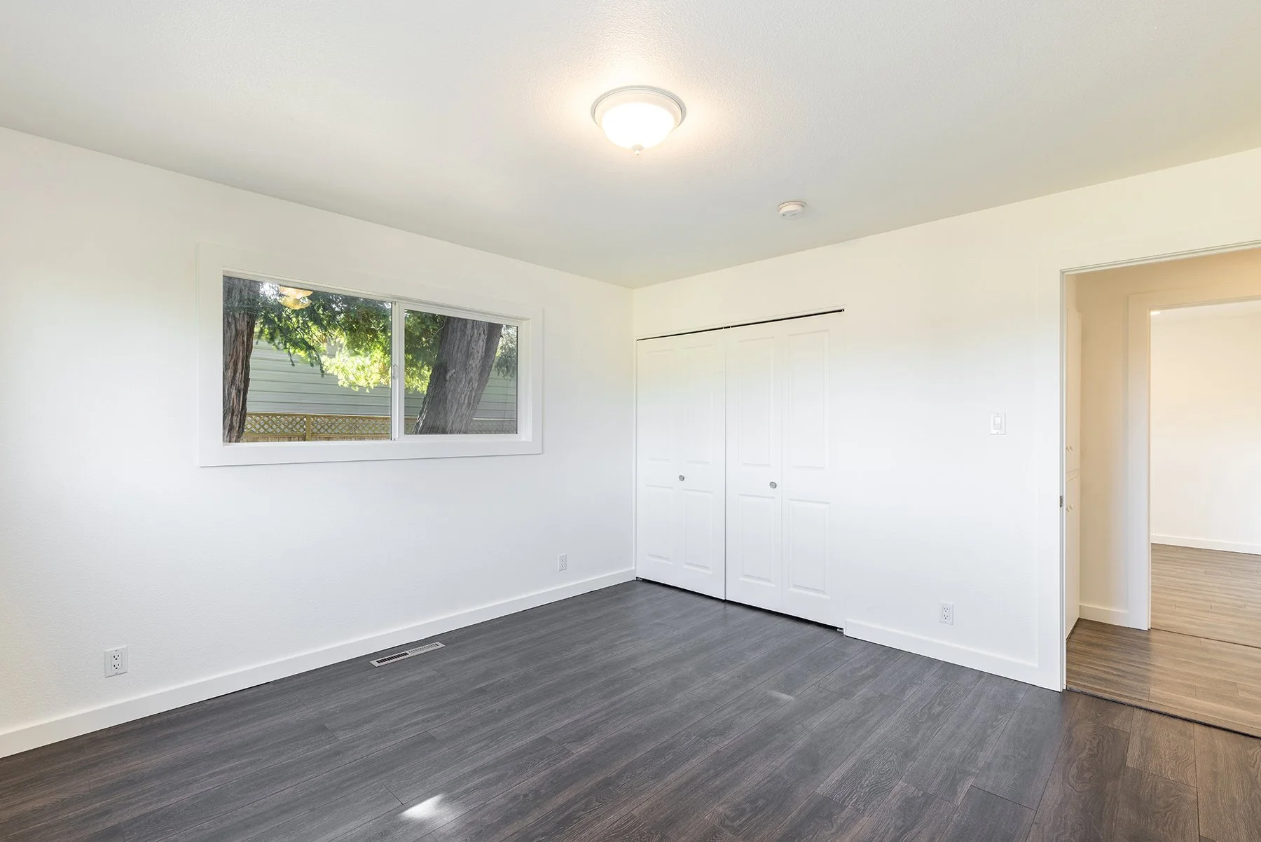 Empty room with white walls, dark wood flooring, a window showing trees outside, and a closet with white double doors.