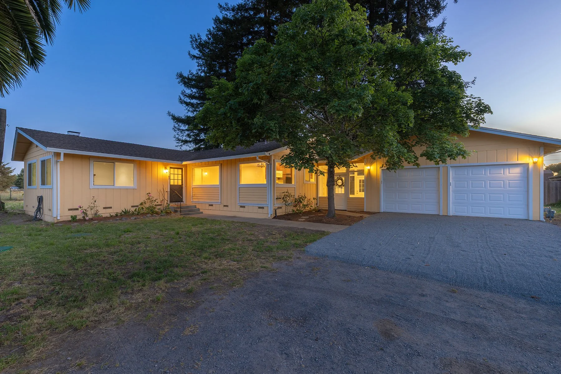 A single-story, yellow house with white trim, a garage, and a large tree in the front yard. The house has warm yellow lighting from the windows and a lit outdoor light by the front door. The driveway is paved, and the front yard has a small garden ar