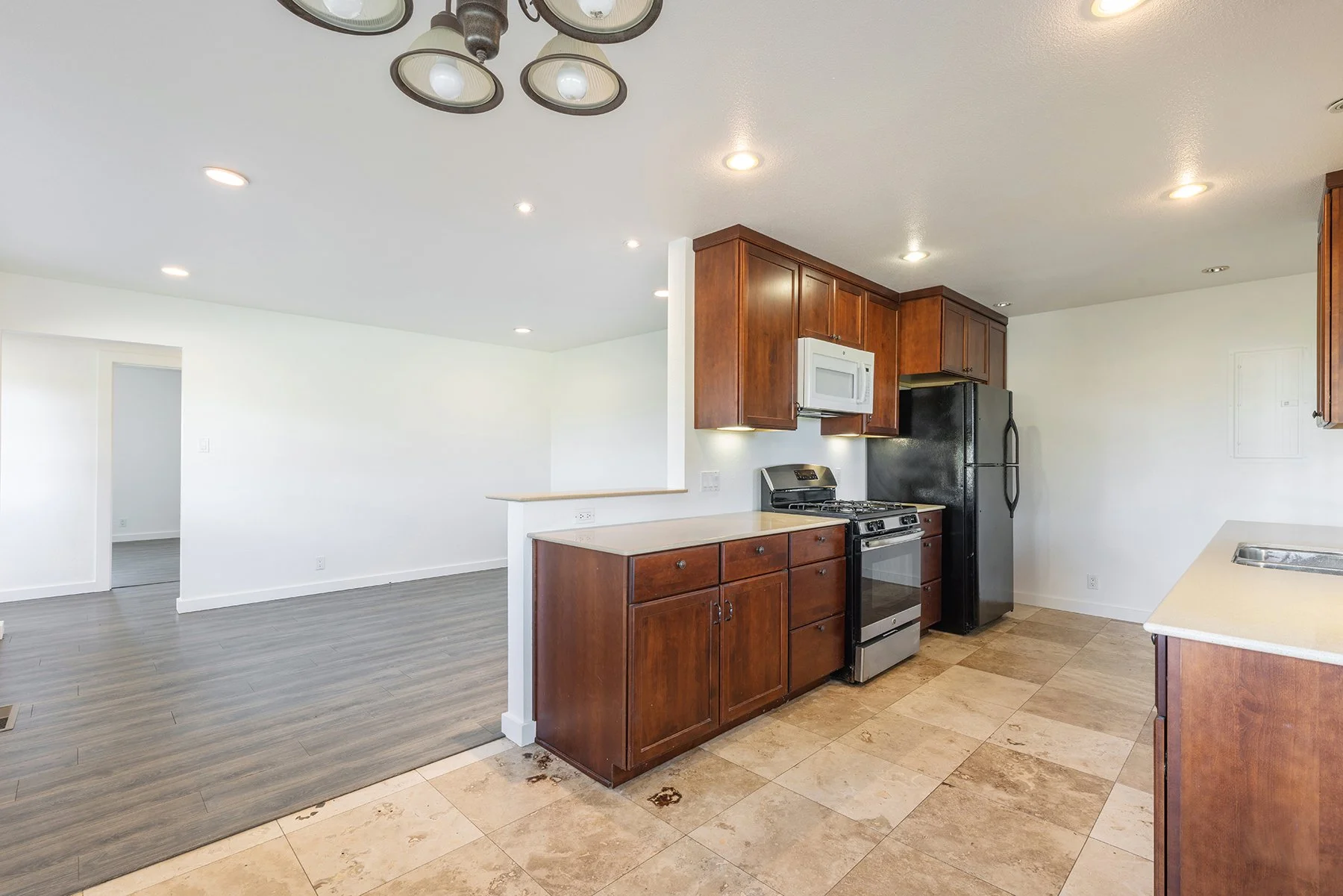 Kitchen area with wood cabinets, black refrigerator, white microwave, stove, and beige countertops. The flooring is a mix of beige tiles and gray wood-like material. Recessed lighting and a ceiling light fixture are visible.